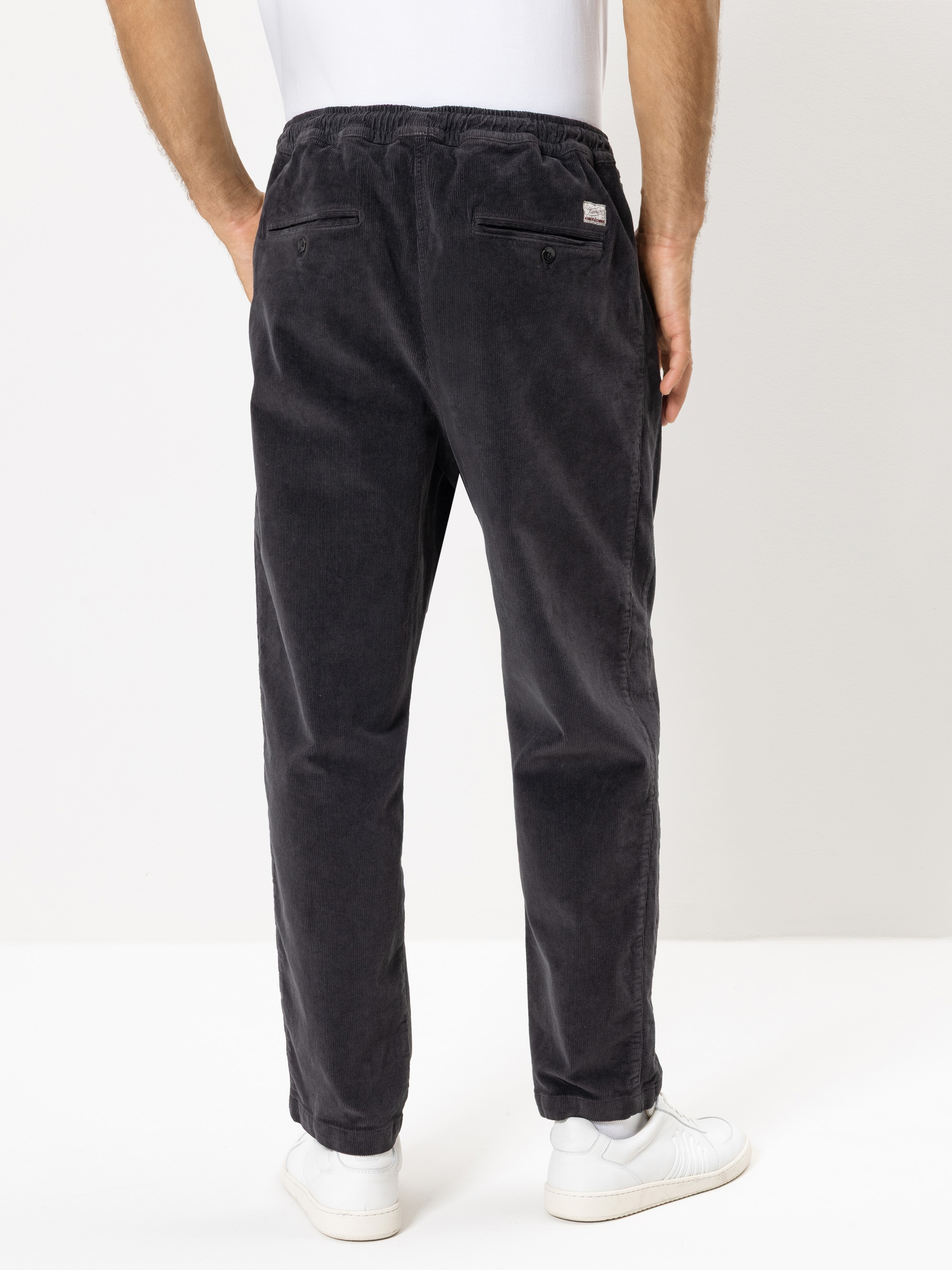 Eine Person, die eine dunkelgraue Cordhose, ein weißes Hemd und weiße Turnschuhe trägt, wird von hinten gezeigt und steht vor einem schlichten hellen Hintergrund.