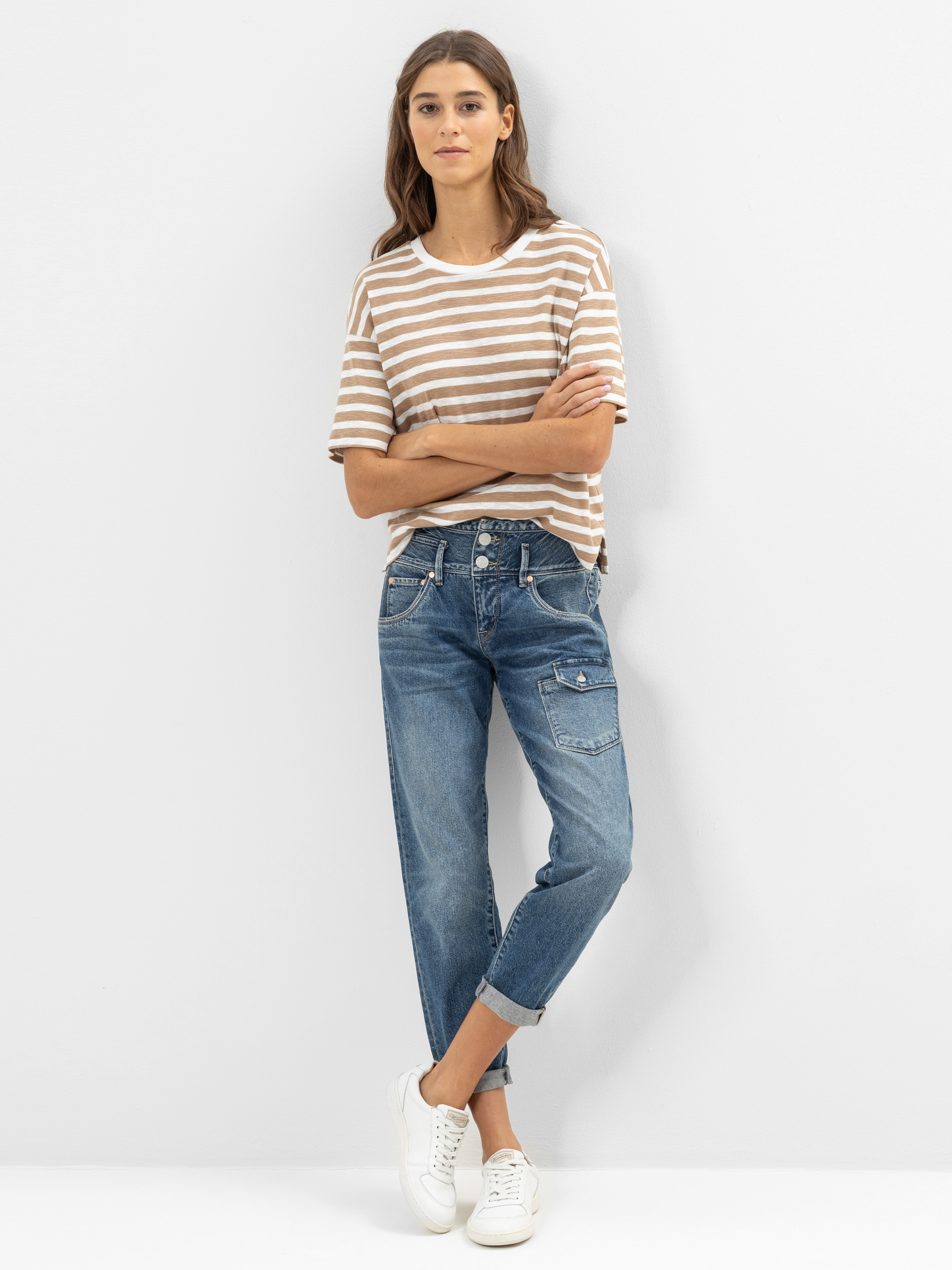 Eine Frau steht an einer weißen Wand und trägt ein braun-weiß gestreiftes T-Shirt, eine Jeans mit blauen Bündchen und Taschen sowie weiße Turnschuhe. Sie hat langes braunes Haar und ihre Arme sind locker verschränkt.