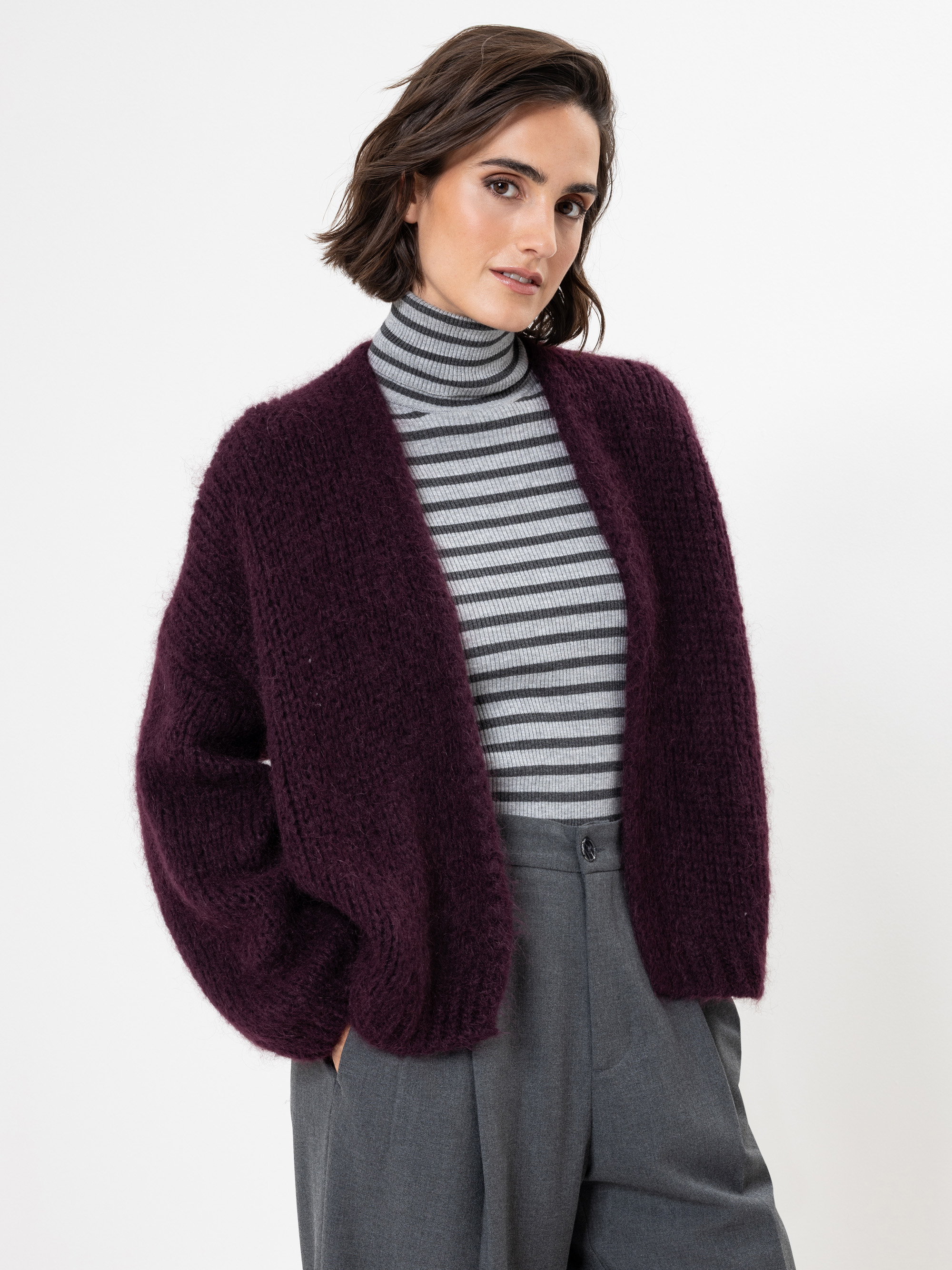Eine Frau mit kurzen braunen Haaren trägt eine gemütliche dunkelviolette Strickjacke über einem grau-weiß gestreiften Rollkragenpullover und eine hochgeschlossene graue Hose vor einem schlichten weißen Hintergrund.