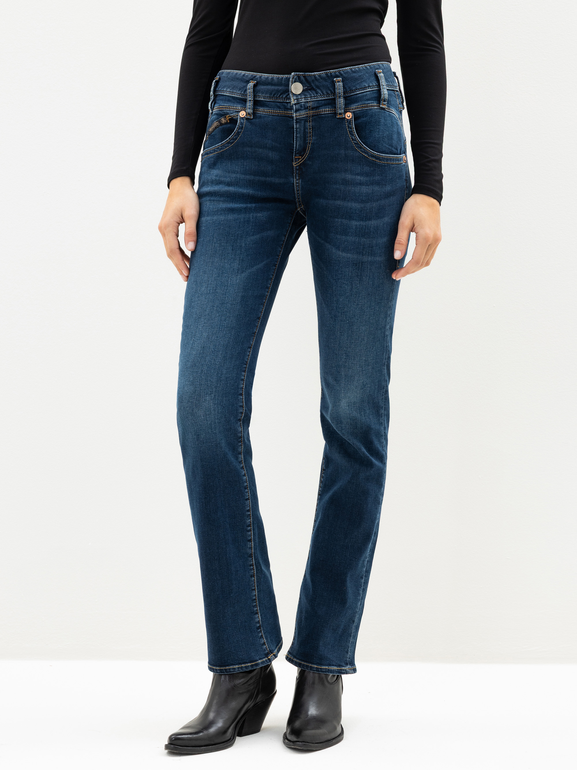 Eine Person, die ein schwarzes langärmeliges Oberteil, dunkelblaue Jeans mit geradem Bein und schwarze Stiefeletten trägt, steht vor einem schlichten weißen Hintergrund.