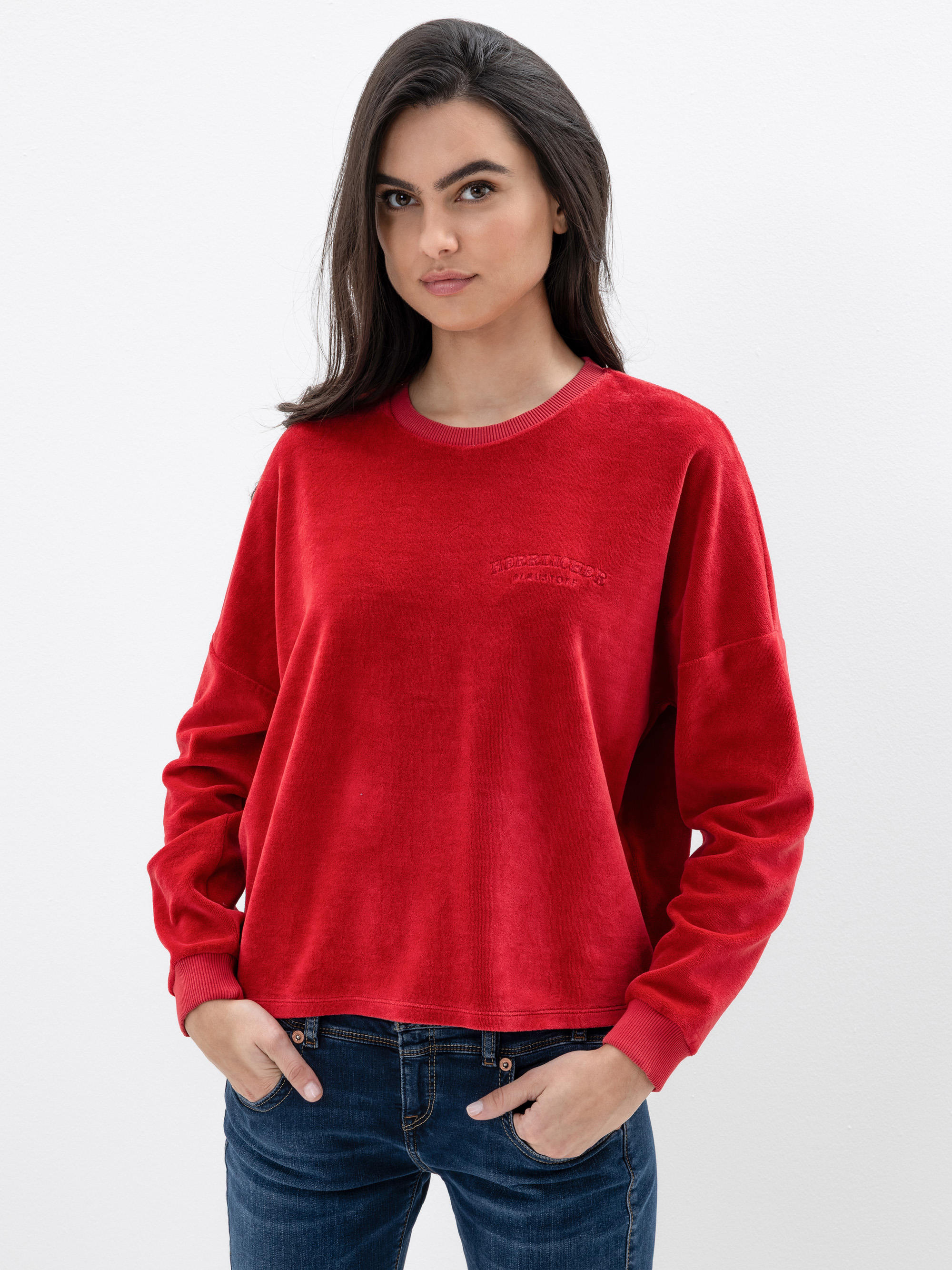 Eine Frau mit langen dunklen Haaren trägt ein locker sitzendes rotes Samtsweatshirt und blaue Jeans und steht mit den Händen in den Taschen vor einem schlichten weißen Hintergrund.