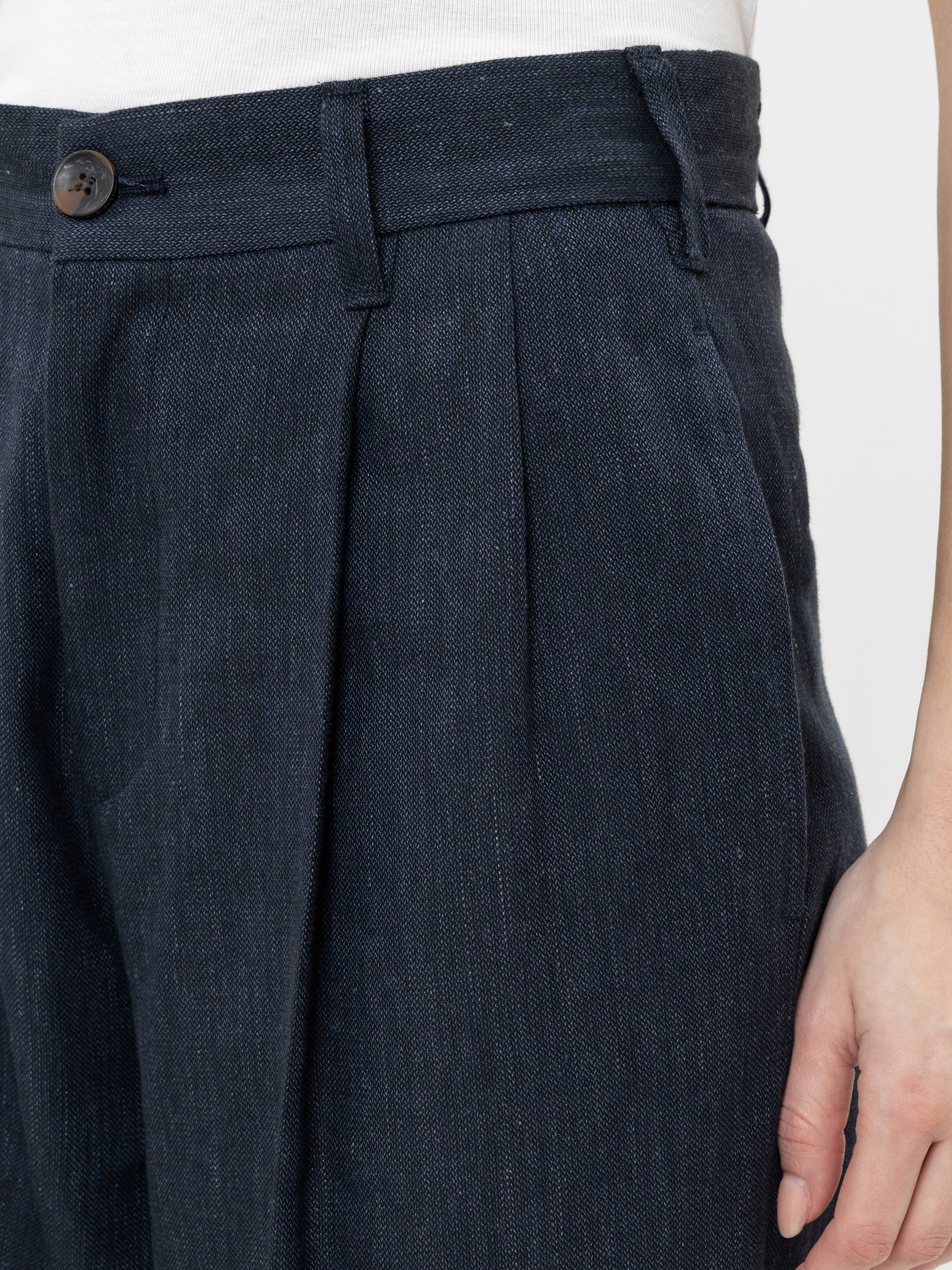 Nahaufnahme einer Person, die eine dunkelblaue Bundfaltenhose mit hoher Taille, Knopfverschluss und Gürtelschlaufen trägt. Die rechte Hand der Person liegt entspannt an ihrer Seite, und ein Teil eines weißen Oberteils ist sichtbar.