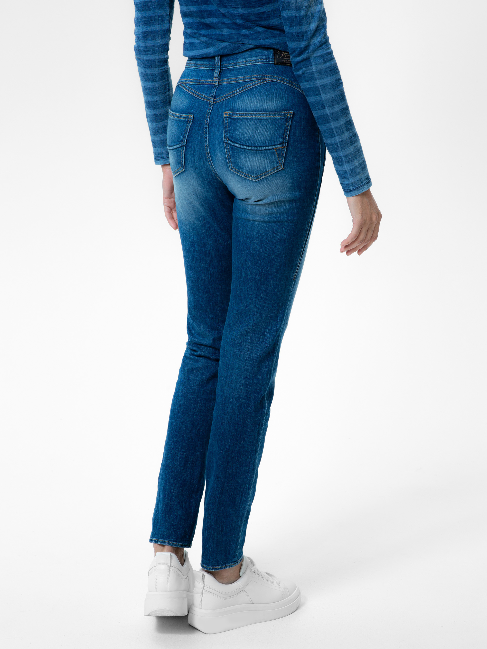 Eine Person, die blaue Skinny-Jeans, ein blau gestreiftes langärmeliges Oberteil und weiße Turnschuhe trägt und mit dem Rücken zur Kamera vor einem schlichten weißen Hintergrund steht.