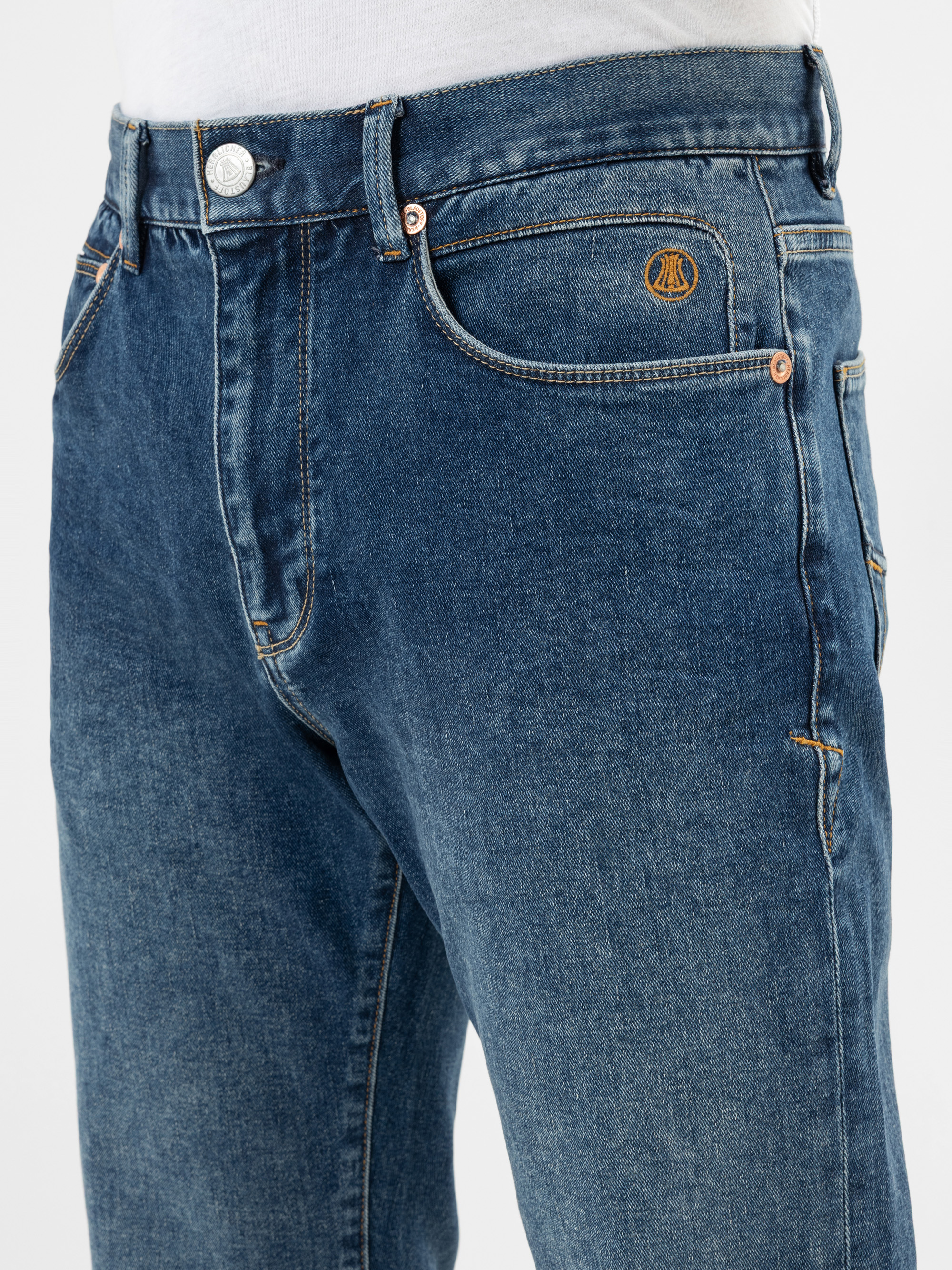 Nahaufnahme einer Person, die eine blaue Jeans mit orangefarbenen Nähten, eine Vordertasche und ein weißes Hemd trägt; über der Tasche ist ein kleines gesticktes Logo zu sehen.