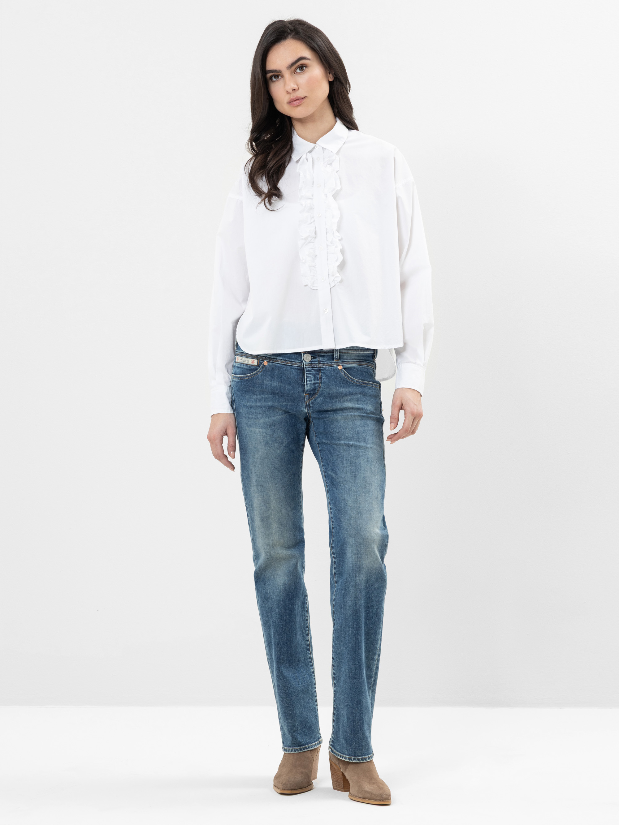 Eine Frau steht vor einem schlichten Hintergrund und trägt eine weiße Rüschenbluse, blaue Jeans und braune Ankle Boots. Sie hat langes dunkles Haar und einen neutralen Gesichtsausdruck, wobei sie die Arme entspannt an der Seite hält.