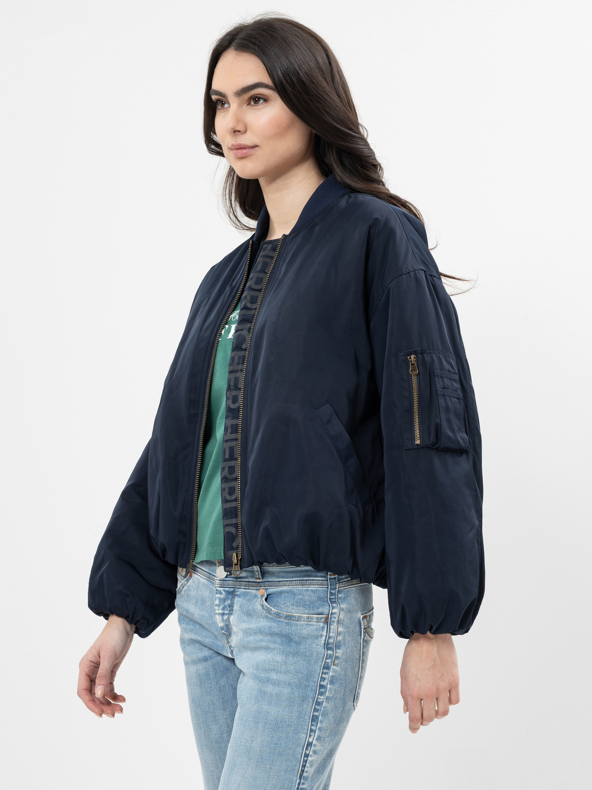 Eine Frau mit langen dunklen Haaren, die eine marineblaue Bomberjacke über einem grünen Oberteil und hellblaue Jeans trägt, steht vor einem schlichten weißen Hintergrund.