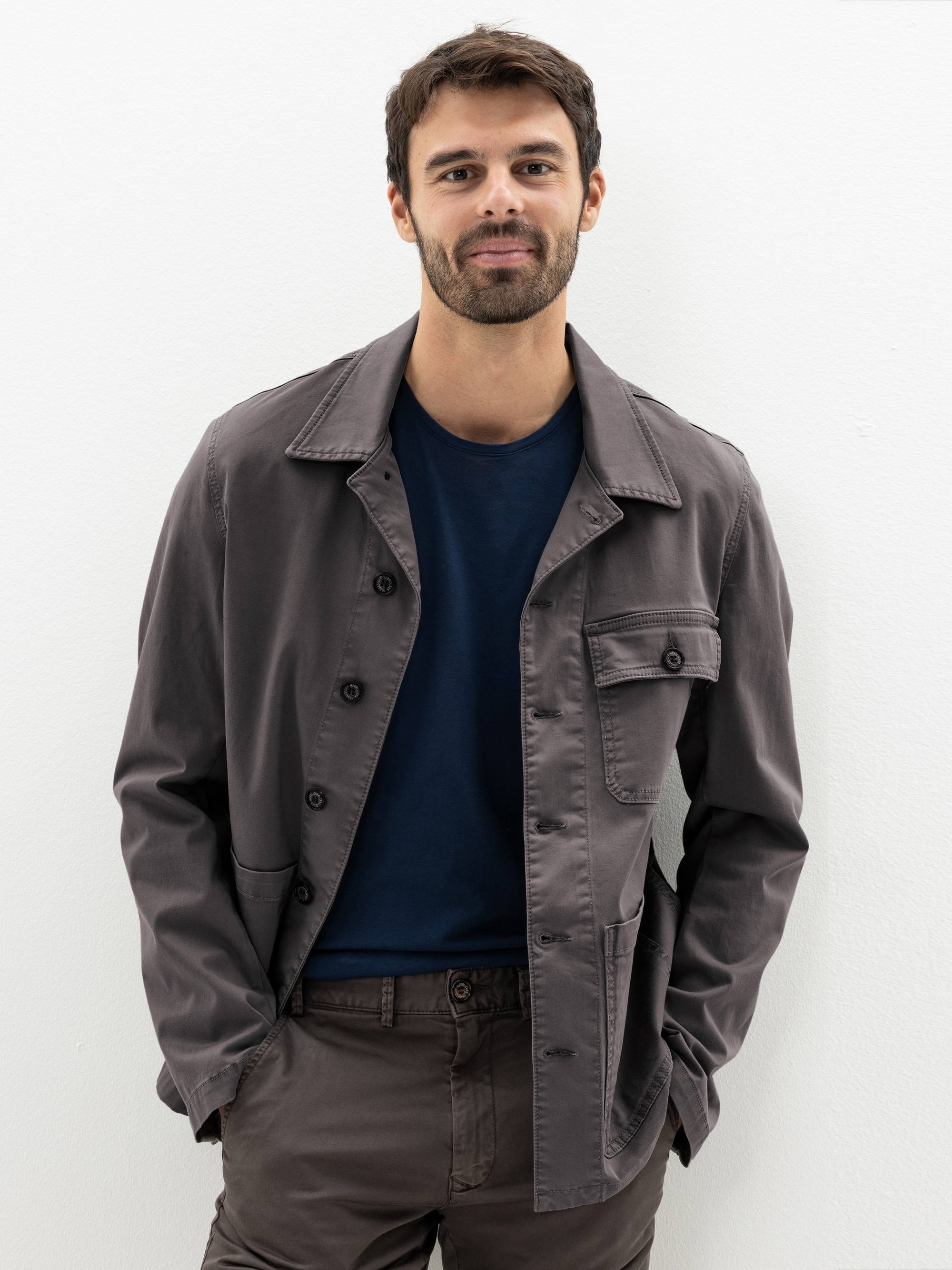 Ein Mann mit kurzen braunen Haaren und Bart steht vor einem weißen Hintergrund, trägt eine graue Jacke über einem marineblauen T-Shirt und eine braune Hose, die Hände in den Taschen und ein leichtes Lächeln im Gesicht.