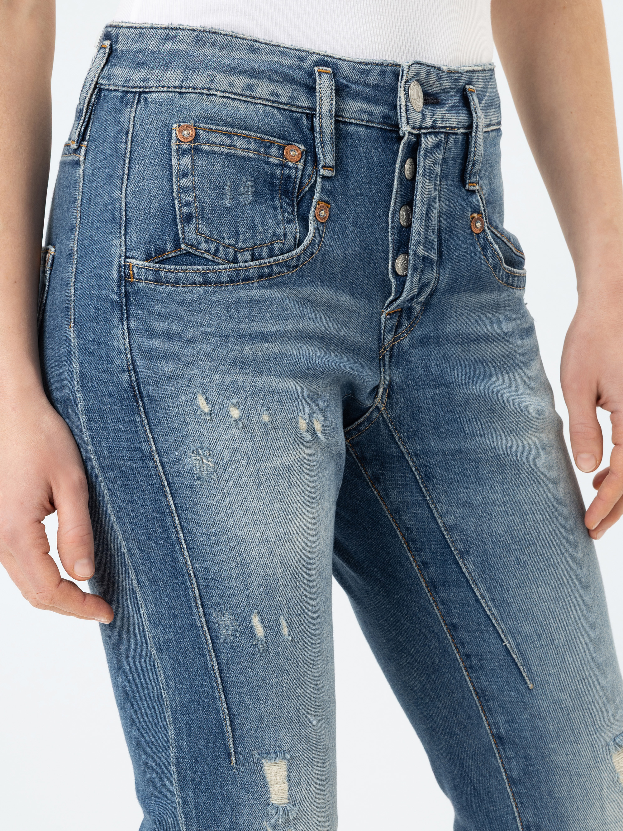 Eine Person trägt eine blaue Jeans mit hoher Taille und abgenutztem Stoff, mit Knopfleiste und Vordertaschen, kombiniert mit einem weißen gerippten Oberteil. Die Jeans hat sichtbare Nähte und abgenutzte Flicken für einen lässigen Look.