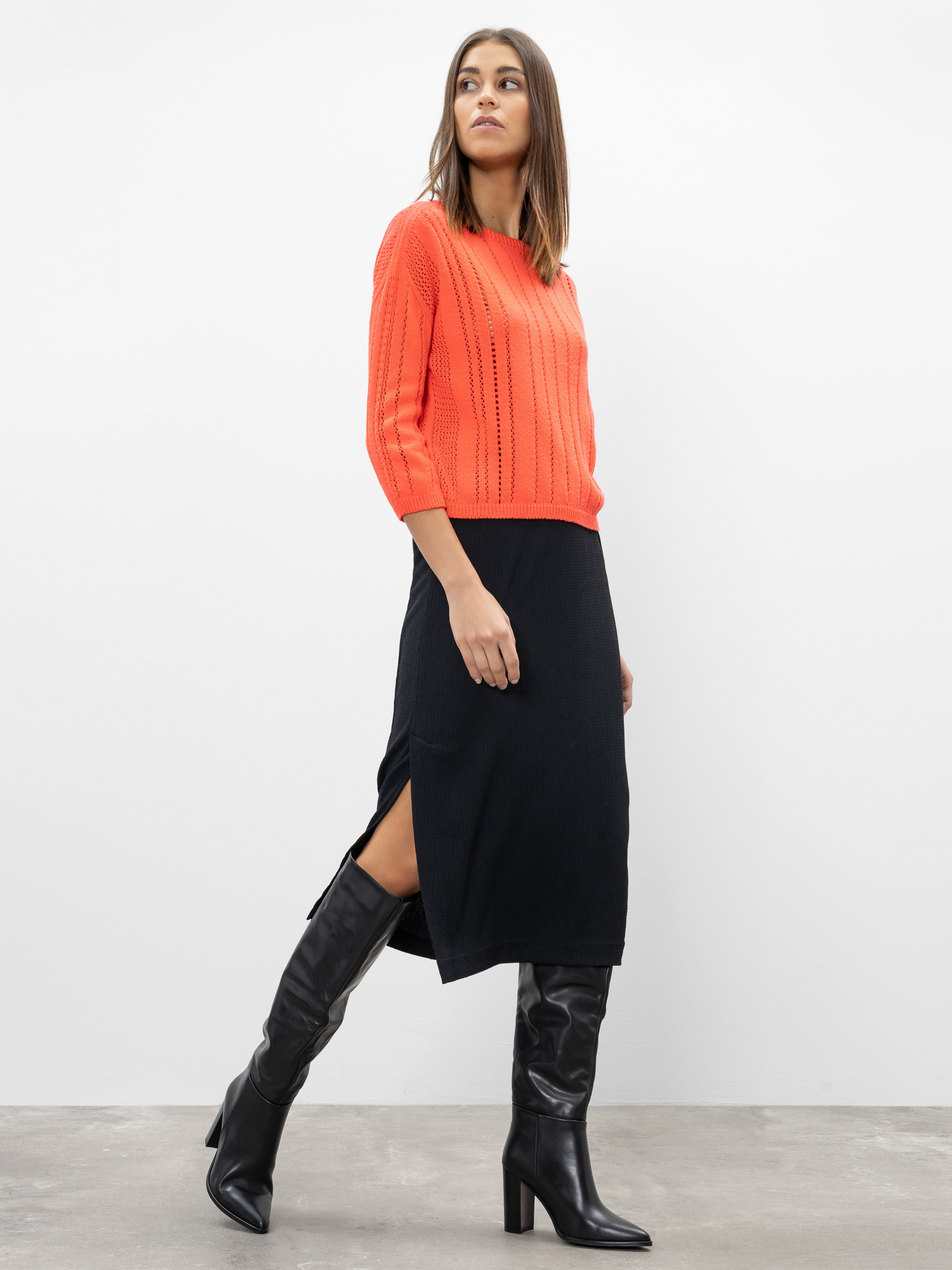Eine Frau steht vor einer schlichten weißen Wand und trägt einen leuchtend orangefarbenen Strickpullover, einen schwarzen Midirock mit Seitenschlitz und kniehohe schwarze Stiefel mit Absatz. Sie blickt mit neutraler Miene zur Seite.