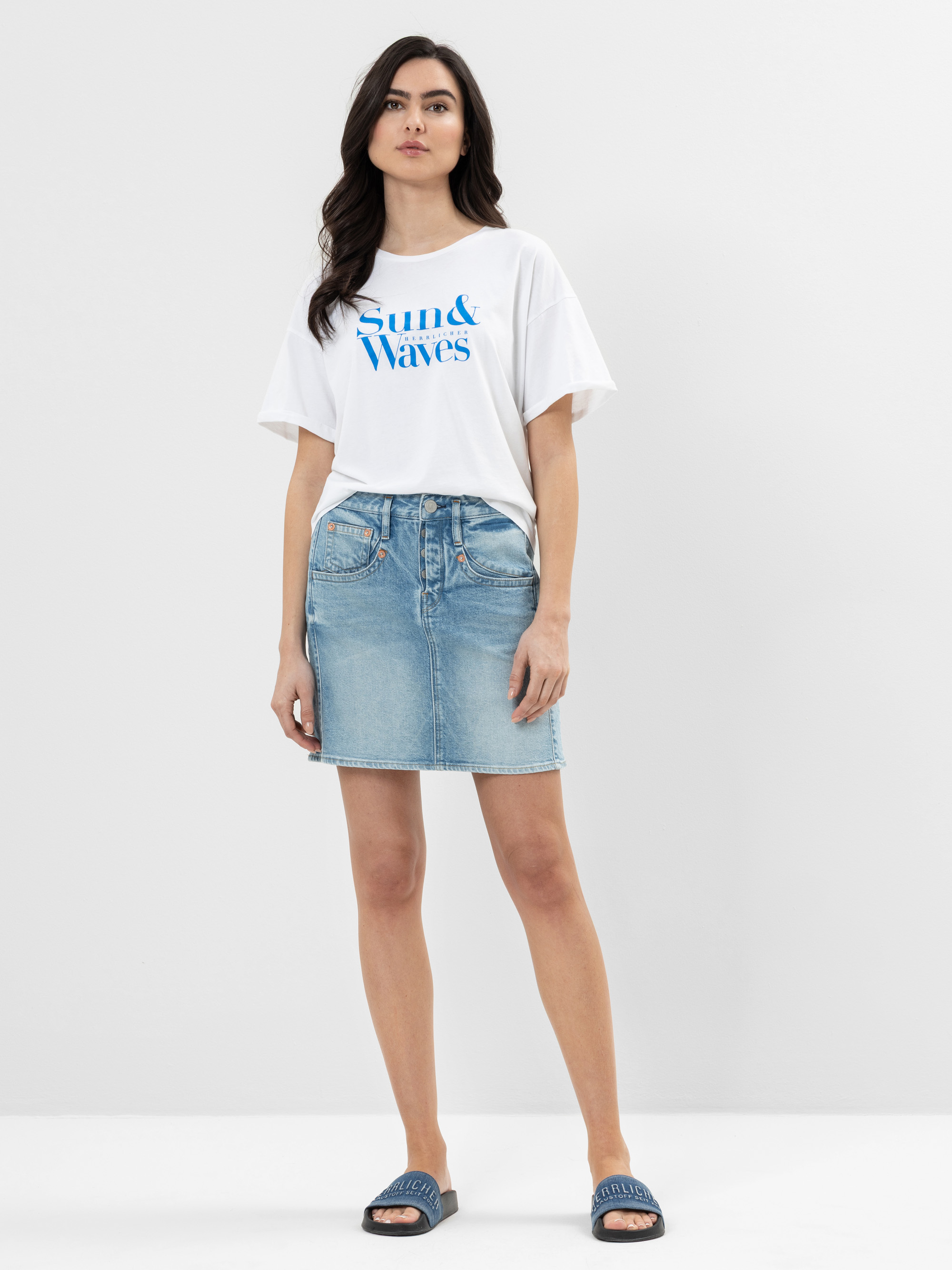 Eine Frau steht vor einem schlichten weißen Hintergrund und trägt ein weißes Sun & Waves-T-Shirt, einen hellblauen Jeansrock und blaue Slide-Sandalen. Sie hat langes dunkles Haar und eine entspannte, selbstbewusste Körperhaltung.