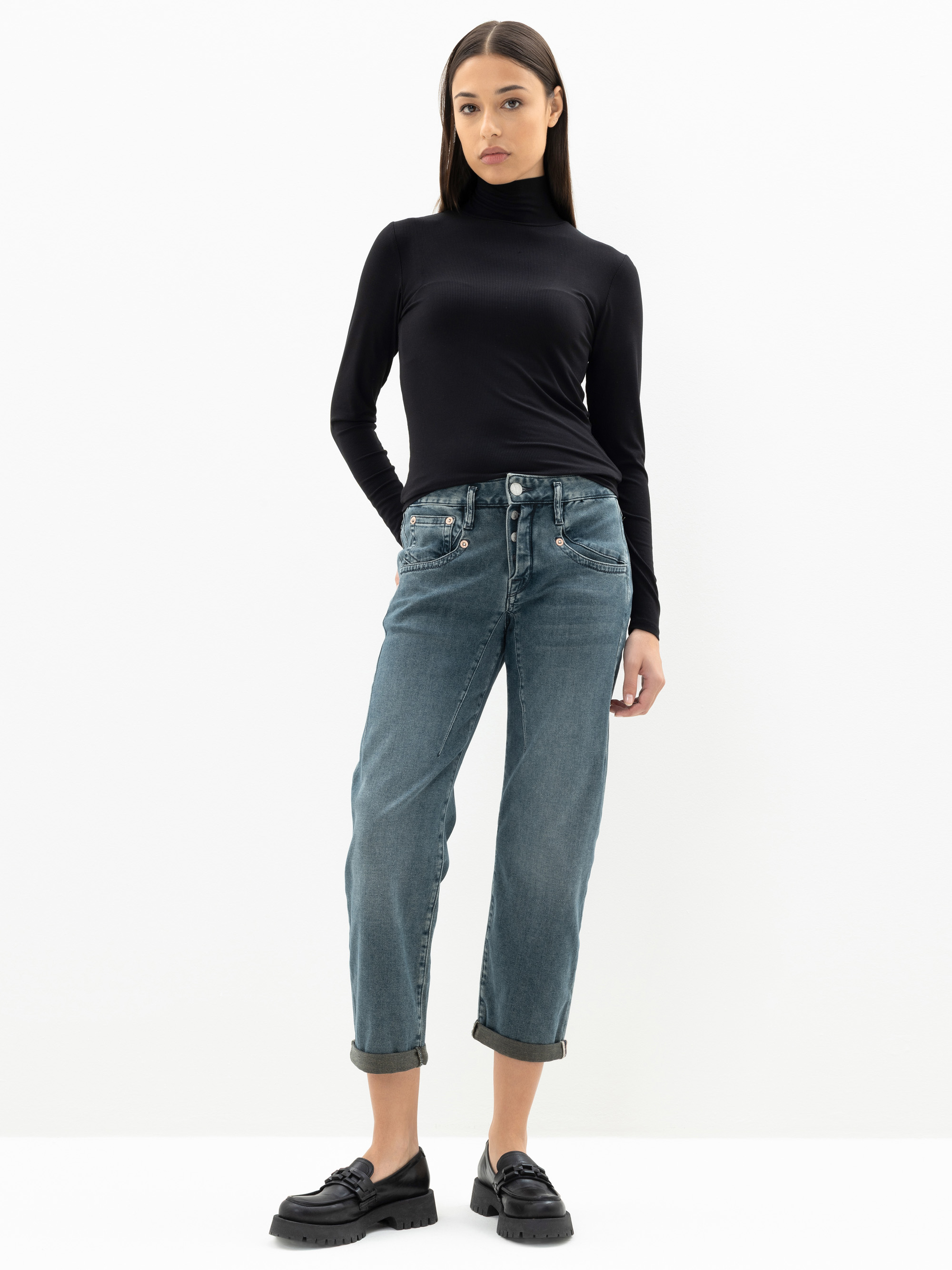 Eine Frau steht vor einem schlichten weißen Hintergrund und trägt einen schwarzen langärmeligen Rollkragenpullover, blaue Jeans mit Bündchen und schwarze Halbschuhe. Sie hat glattes dunkles Haar und einen neutralen Gesichtsausdruck.