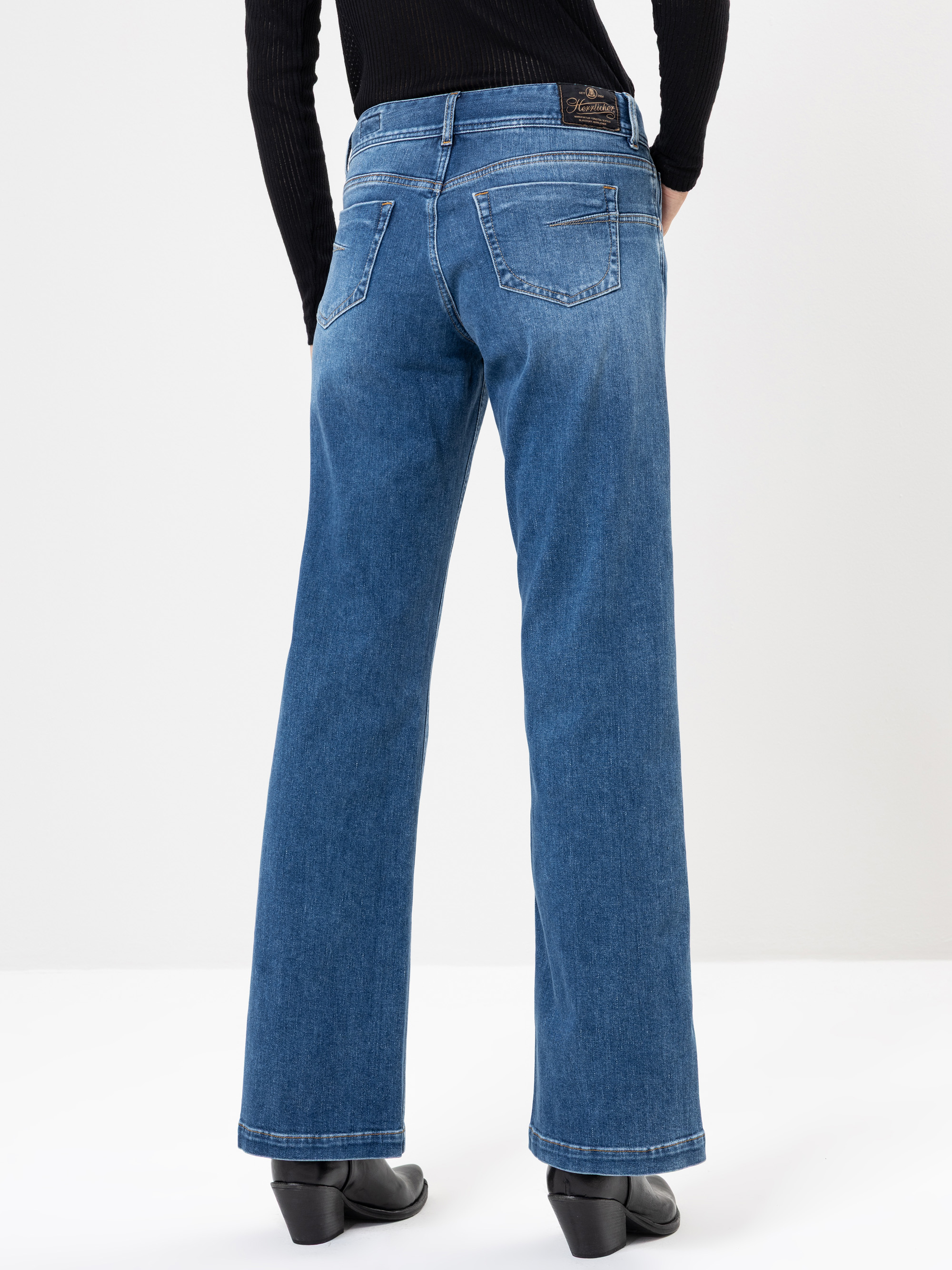 Eine Person, die eine blaue Jeans mit weitem Bein, ein schwarzes langärmeliges Oberteil und schwarze Schuhe trägt, wird von hinten vor einem schlichten weißen Hintergrund gezeigt.