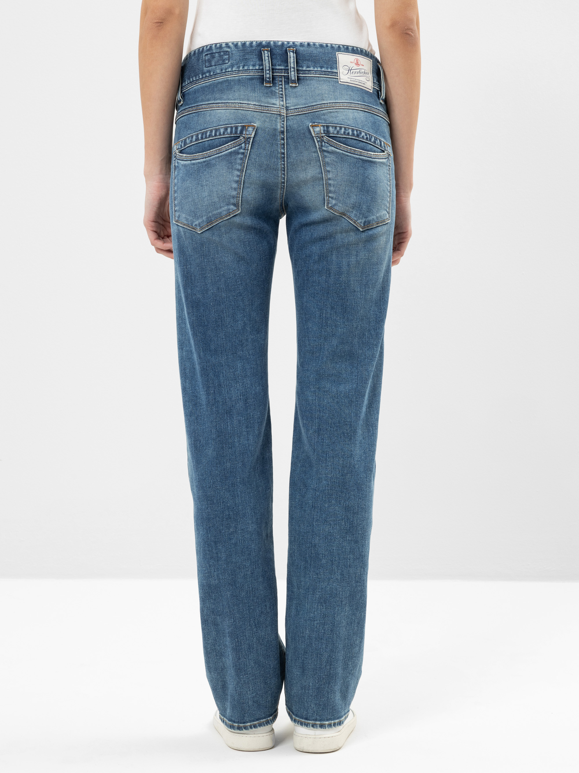 Person, die mit dem Gesicht zur Wand steht und eine blaue Jeans mit geradem Bein, sichtbaren Gesäßtaschen, einem weißen Etikett am Bund und weißen Schuhen vor einem schlichten hellen Hintergrund trägt.