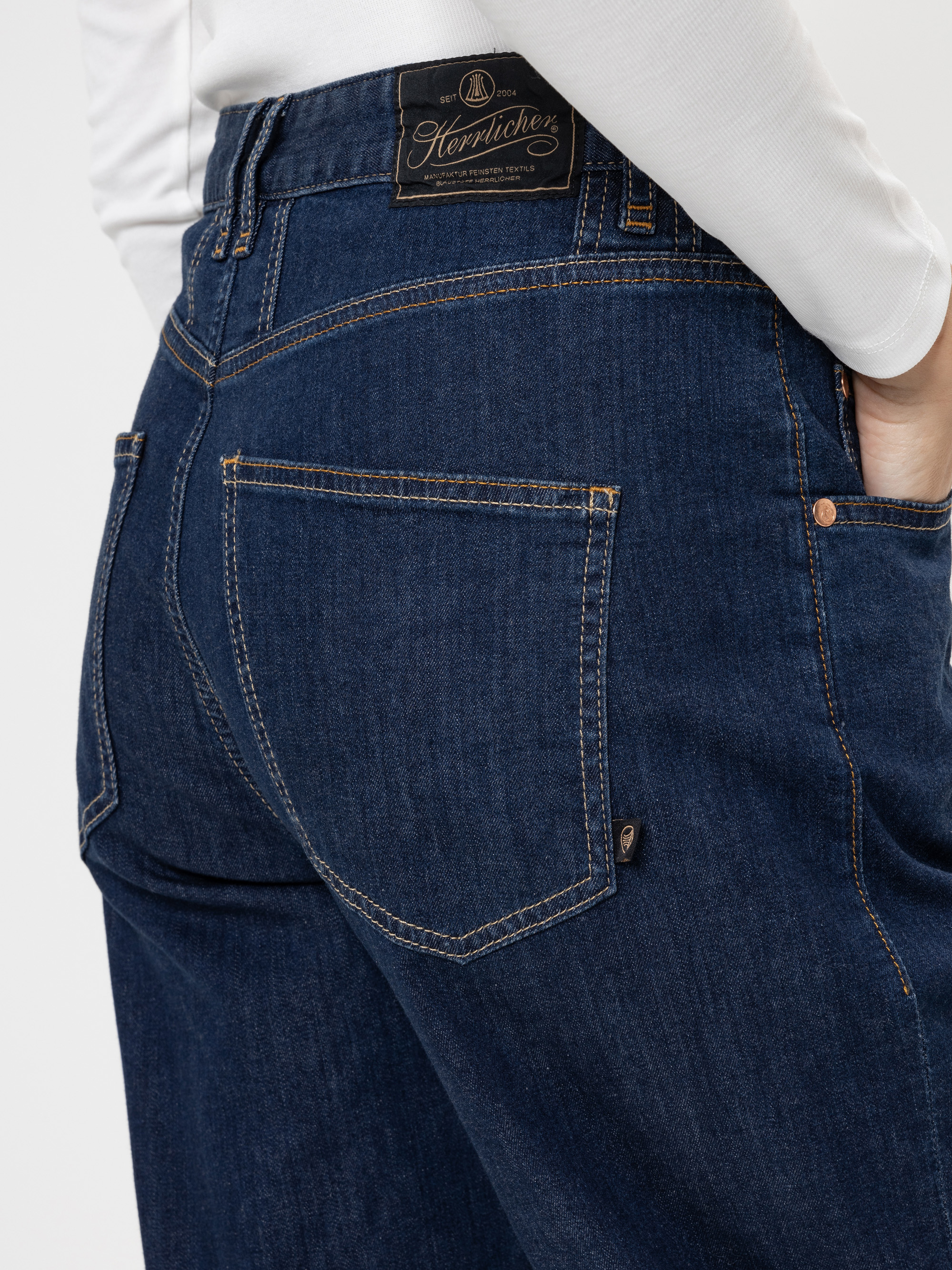 Nahaufnahme einer Person, die eine dunkelblaue Denim-Jeans mit sichtbaren Nähten und einem schwarzen Markenaufnäher aus Leder am Bund trägt, wobei die Gesäßtasche und ein Teil eines weißen Oberteils zu sehen sind.