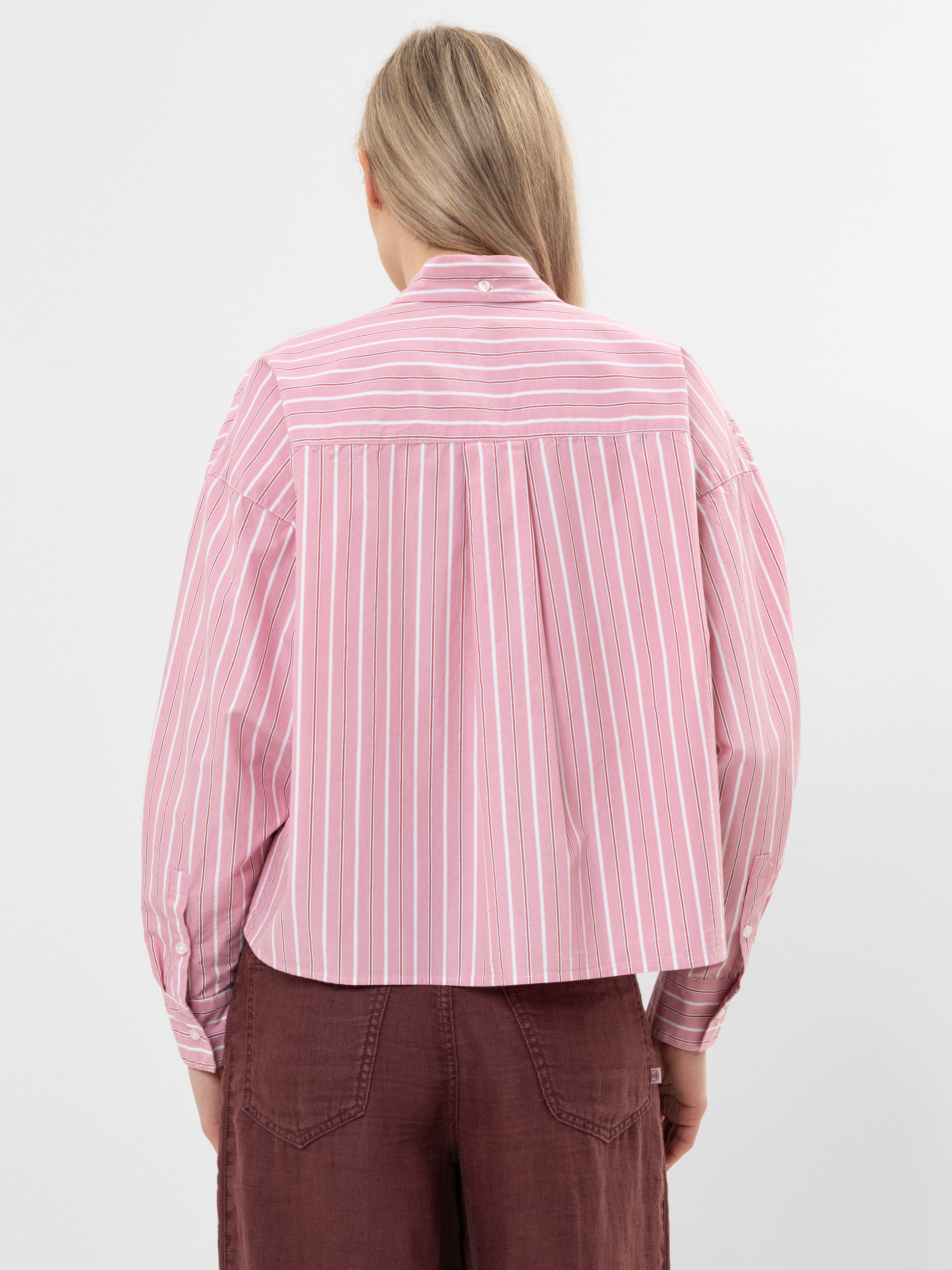 Eine Person mit langen blonden Haaren ist von hinten zu sehen. Sie trägt ein rosa-weiß gestreiftes, langärmeliges Hemd mit Kragen und lockerem Sitz, gepaart mit einer braunen Hose mit hoher Taille.