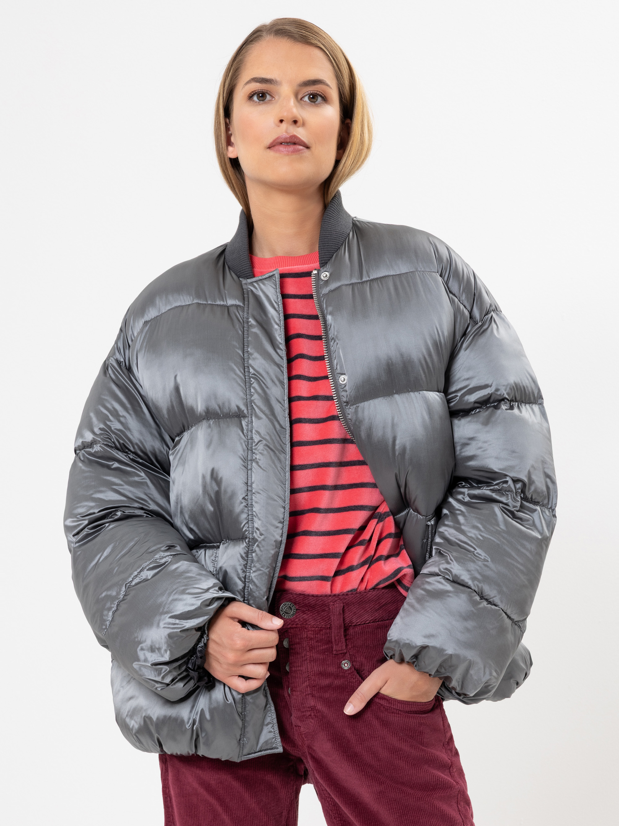 Eine Person, die eine glänzende, silberne Pufferjacke über einem rot-rosa gestreiften Hemd und einer kastanienbraunen Cordhose trägt, steht vor einem schlichten weißen Hintergrund und schaut selbstbewusst in die Kamera.