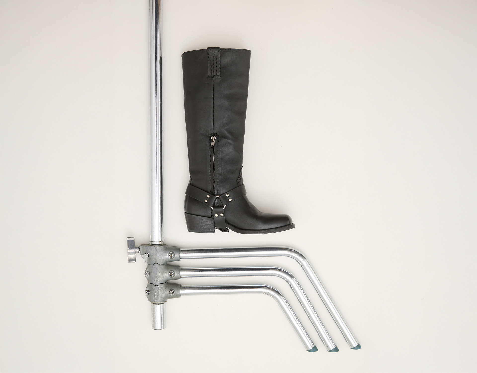 Ein schwarzer kniehoher Stiefel mit Schnallen ist horizontal auf drei gebogenen Metallstangen positioniert, die an einem vertikalen Metallständer befestigt sind, alles vor einem schlichten hellen Hintergrund.