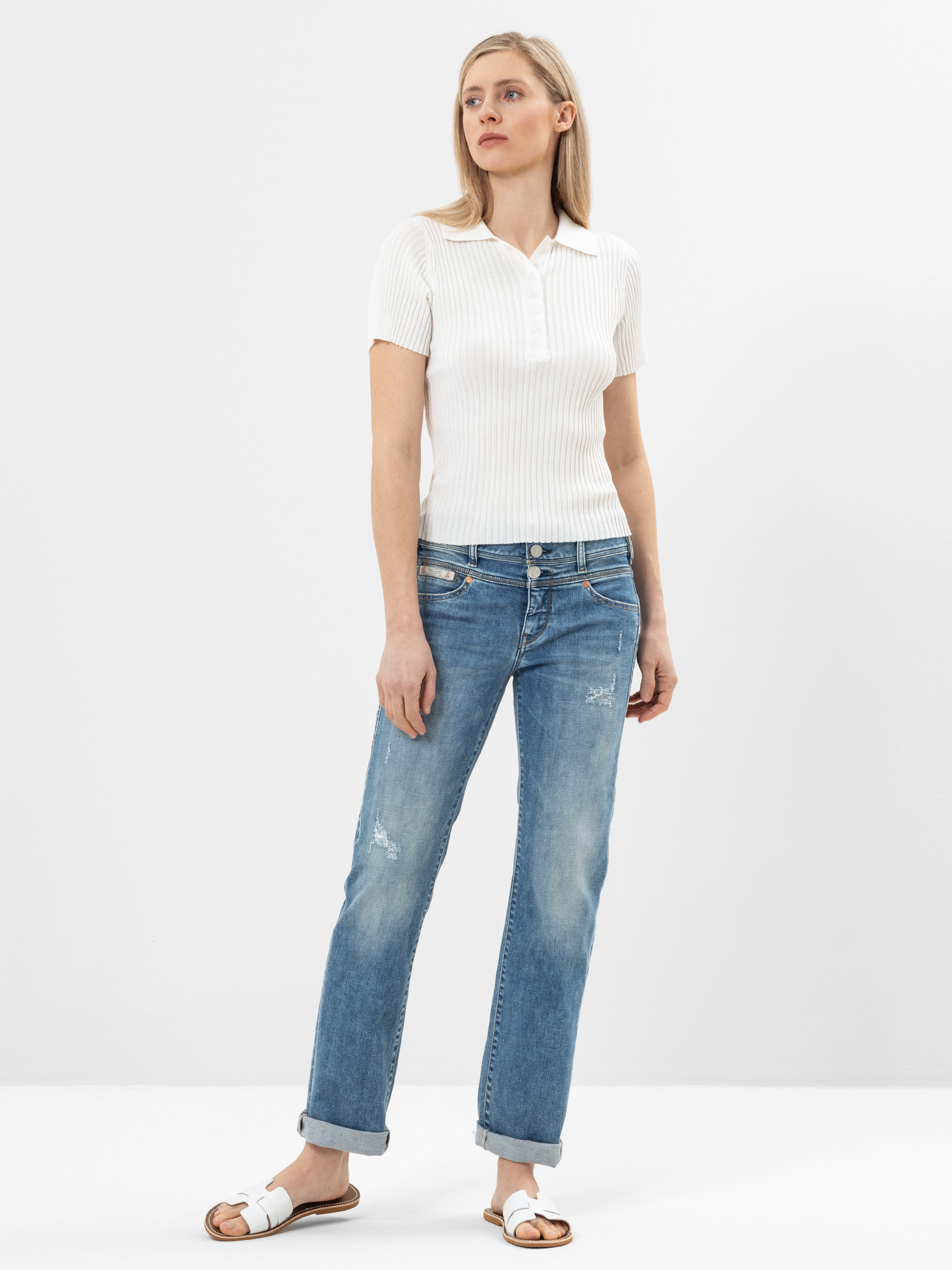 Eine Frau steht vor einem schlichten weißen Hintergrund und trägt ein kurzärmeliges, weißes, geripptes Poloshirt, hellblaue, locker sitzende Jeans und weiße Slide-Sandalen. Sie blickt leicht zur Seite und hat einen neutralen Ausdruck.