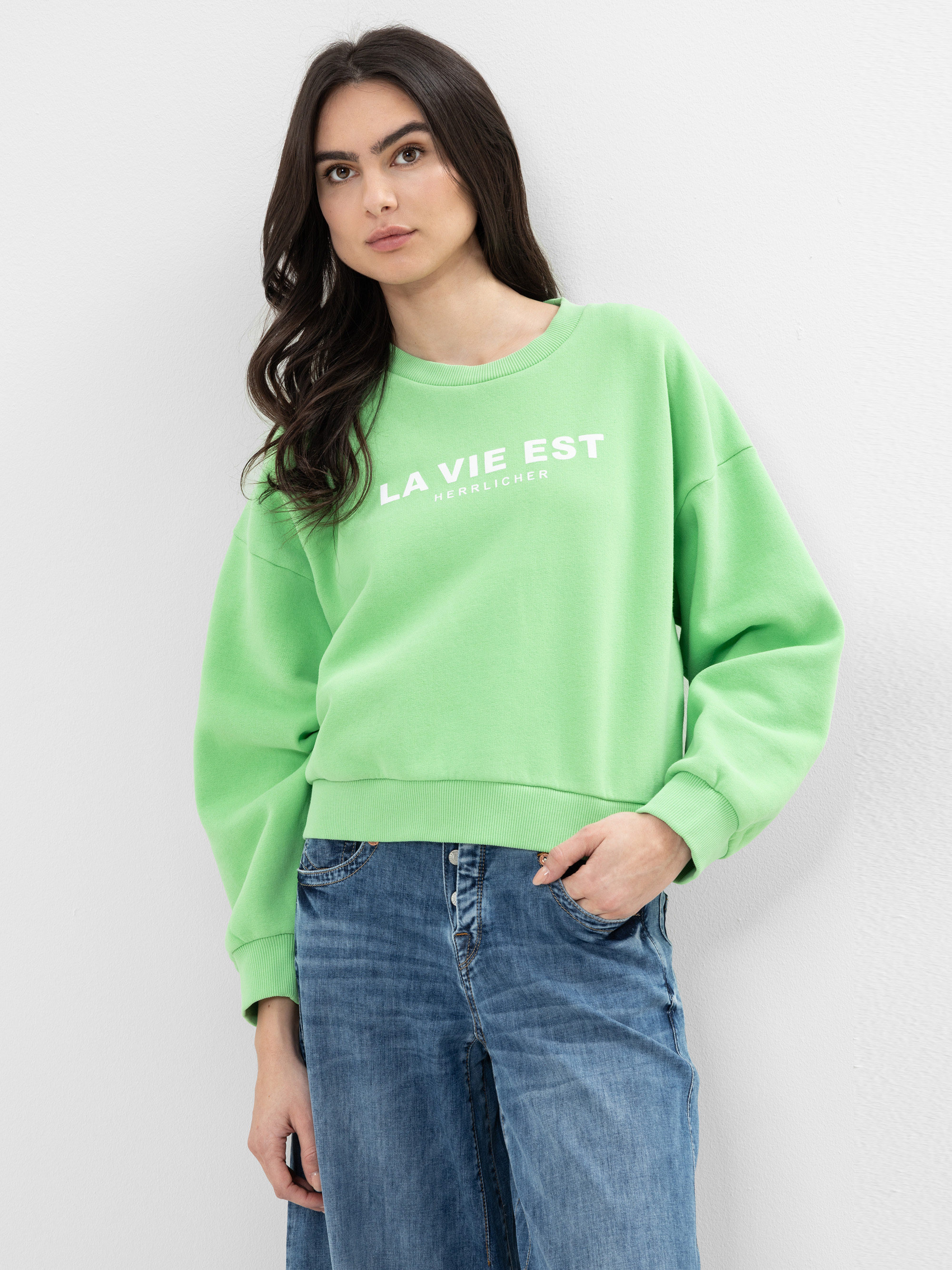 Eine Frau mit langen dunklen Haaren trägt ein hellgrünes Sweatshirt mit dem weißen Aufdruck LA VIE EST, dazu eine blaue Jeans. Sie steht vor einer schlichten weißen Wand und blickt leicht zur Seite.