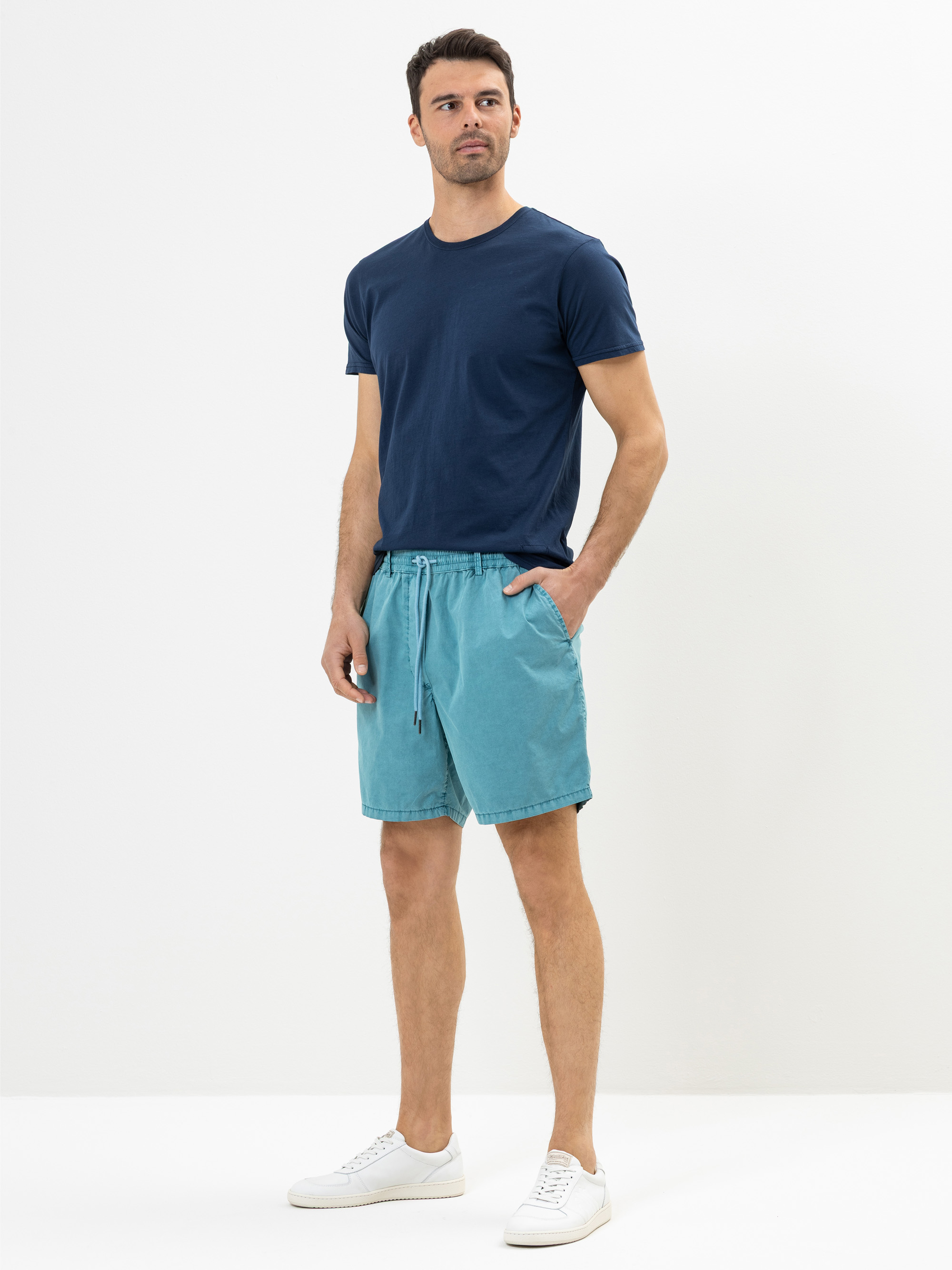 Ein Mann steht vor einem schlichten weißen Hintergrund, trägt ein marineblaues T-Shirt, hellblaue Shorts und weiße Turnschuhe, hat eine Hand in der Tasche und einen neutralen Gesichtsausdruck.
