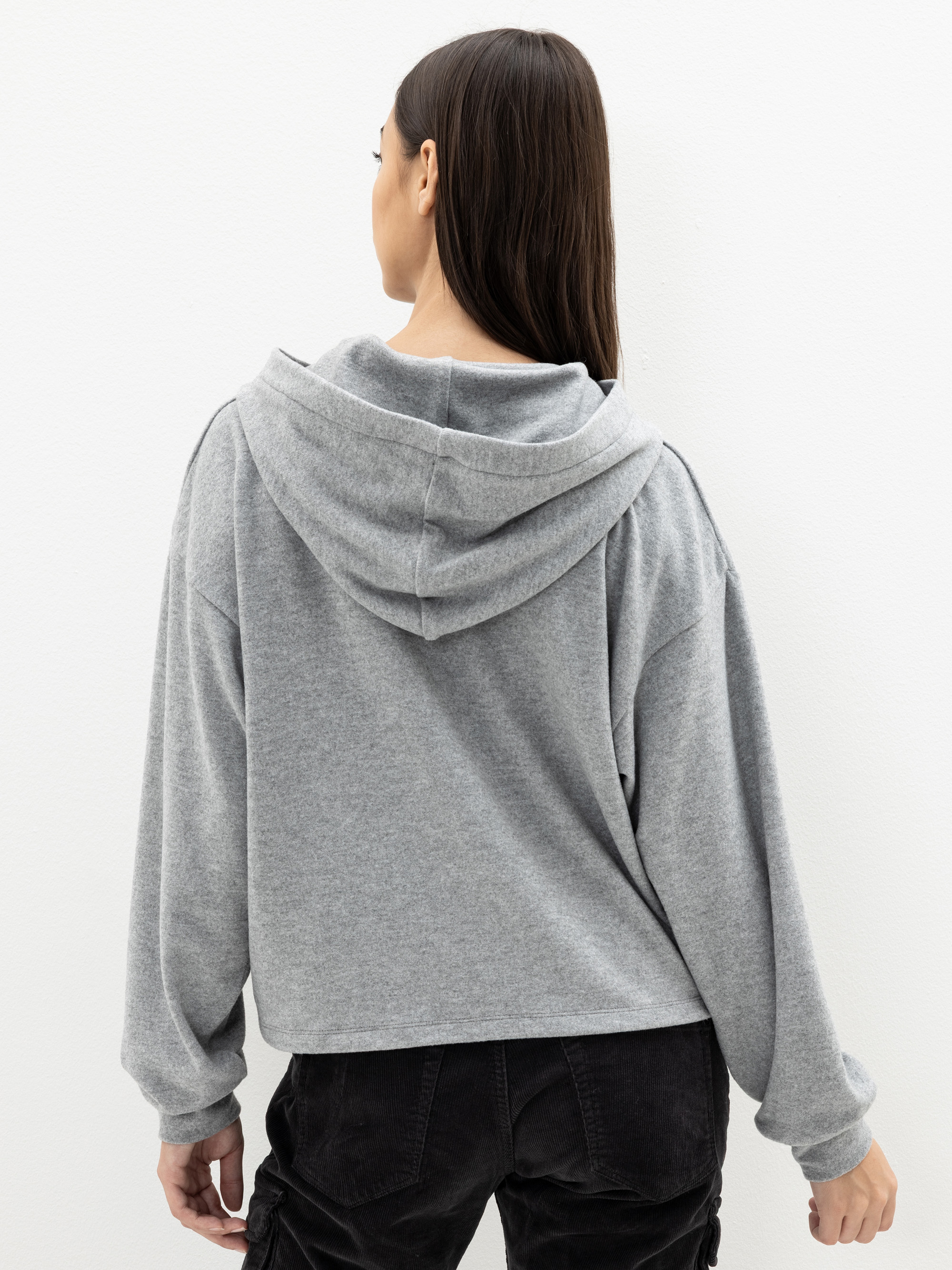 Eine Frau mit langen braunen Haaren steht mit dem Gesicht zur Wand und trägt einen weiten grauen Kapuzenpullover und eine schwarze Hose vor einem schlichten weißen Hintergrund.