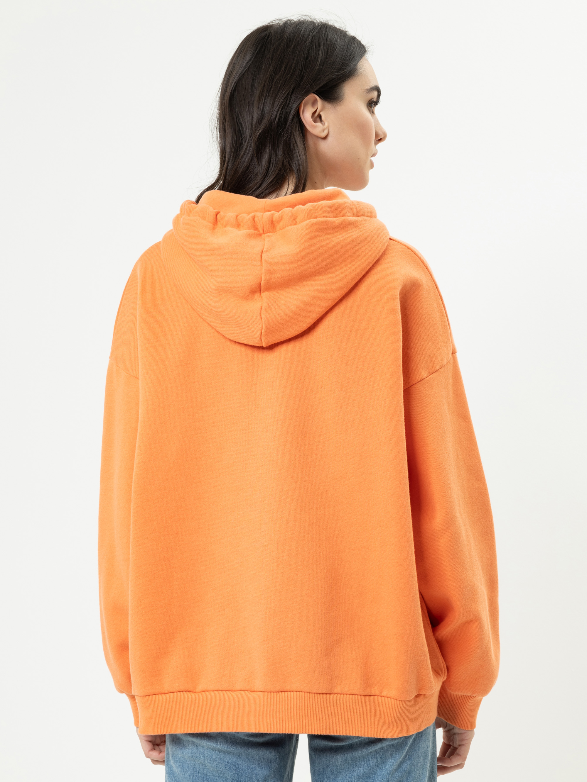 Eine Frau mit dunklen Haaren, die mit dem Rücken zur Kamera steht, trägt einen übergroßen orangefarbenen Kapuzenpullover und blaue Jeans vor einem schlichten weißen Hintergrund.