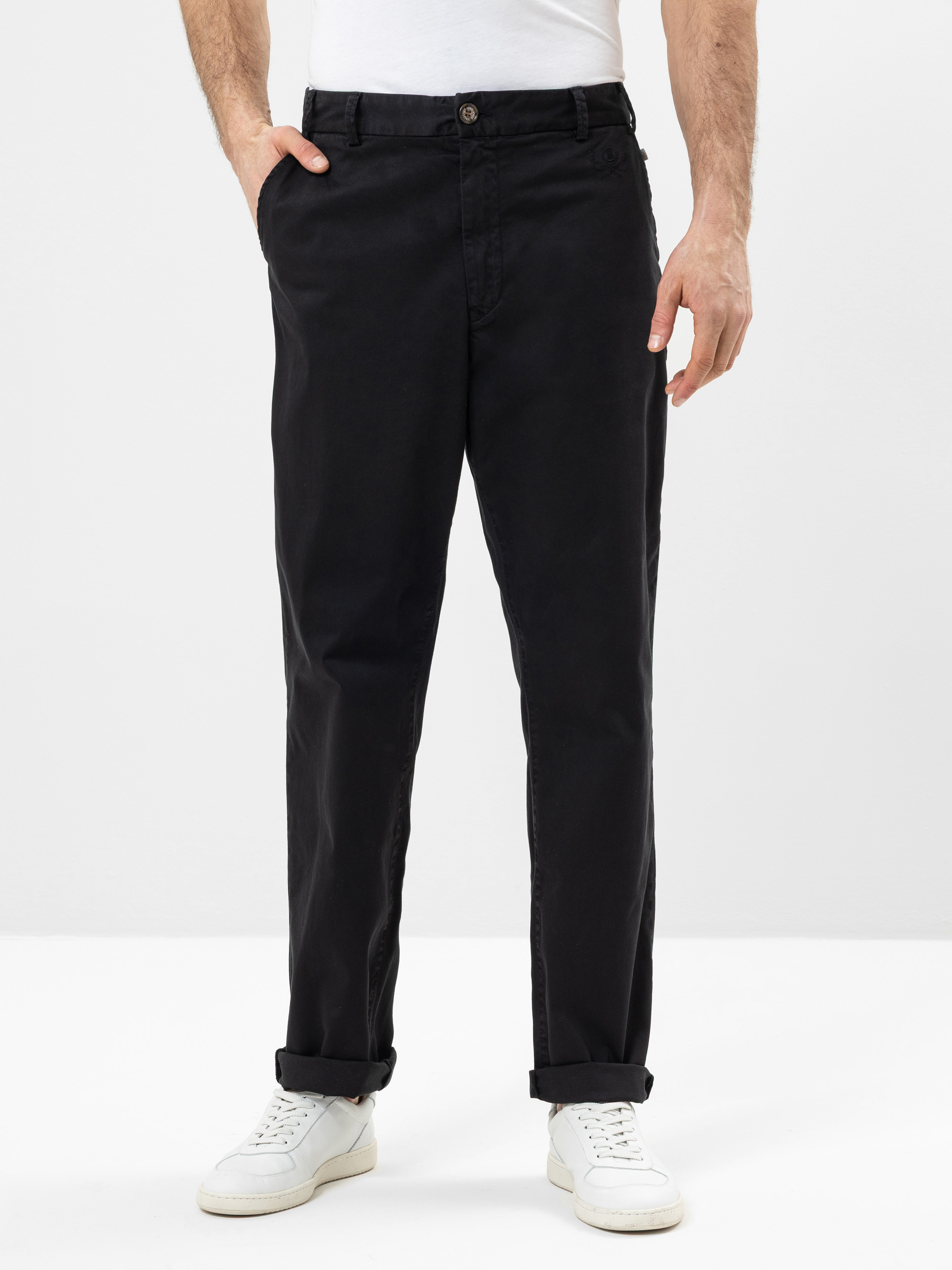 Eine Person, die eine schwarze Hose, ein weißes Hemd und weiße Turnschuhe trägt, steht mit den Händen in den Taschen vor einem schlichten hellen Hintergrund. Das Bild ist von den Schultern abwärts beschnitten.