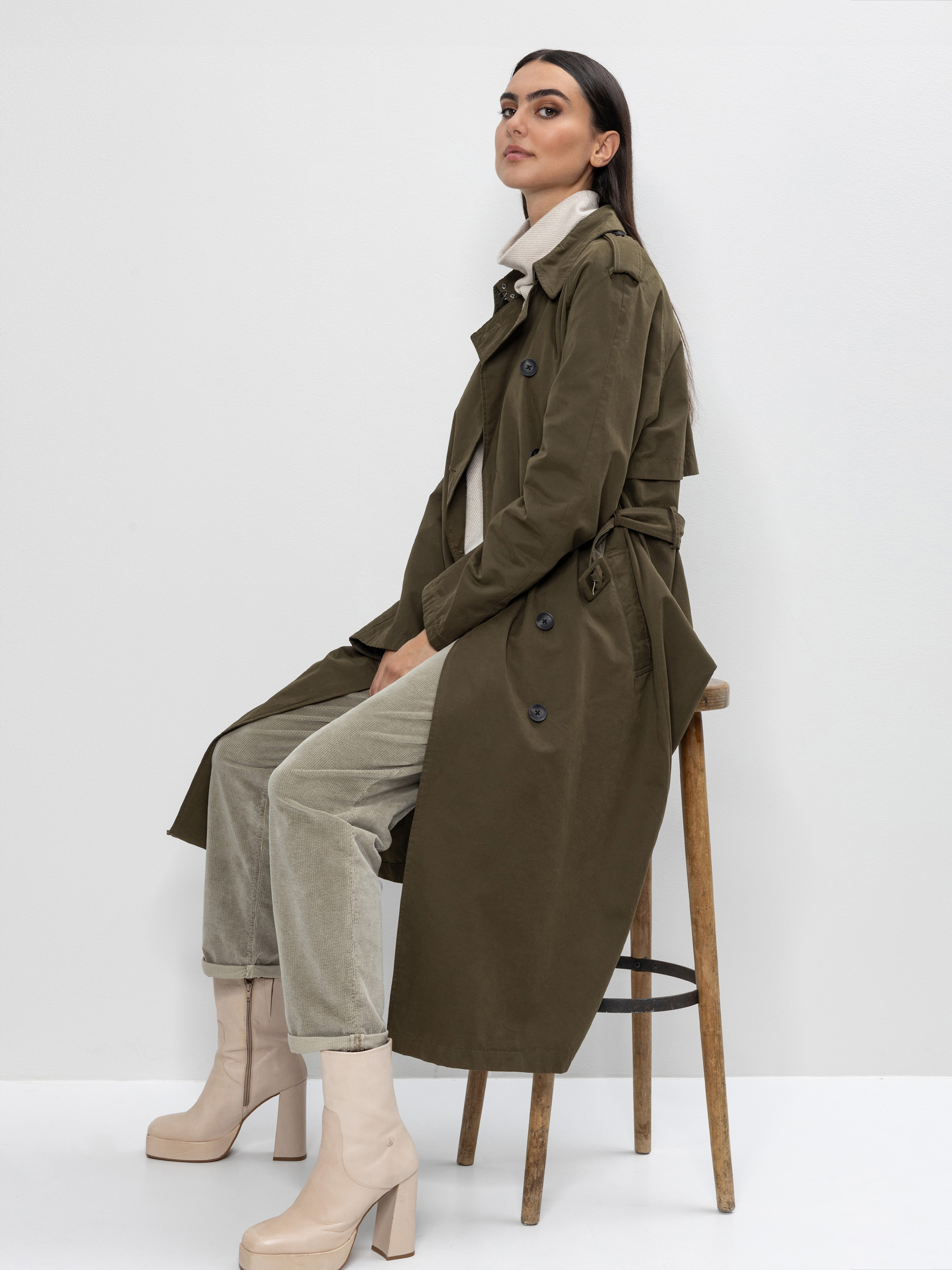 Eine Frau mit langen dunklen Haaren sitzt seitlich auf einem Holzschemel und trägt einen langen olivgrünen Trenchcoat, einen hellen Rollkragenpullover, eine beigefarbene Hose und klobige beigefarbene Stiefel mit Absatz vor einem schlichten weißen Hinte