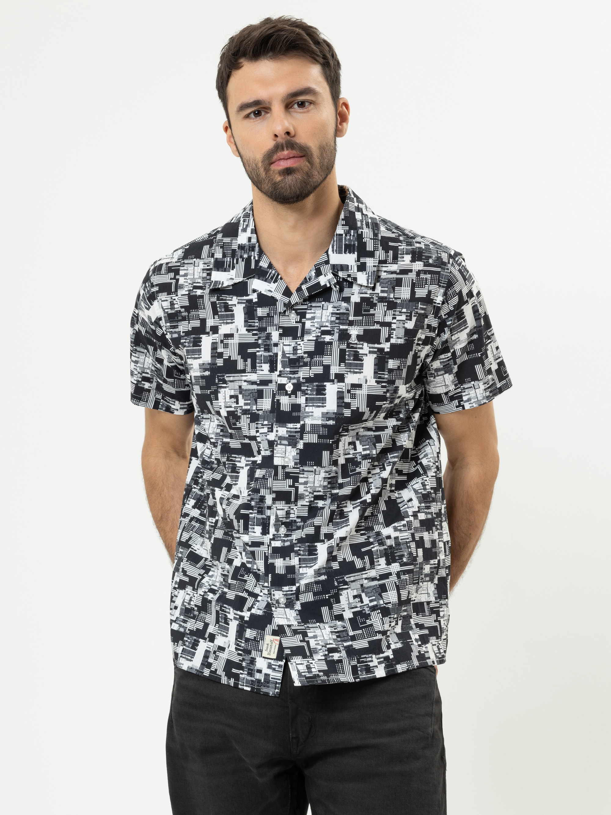 Ein Mann mit kurzen dunklen Haaren und einem Bart trägt ein schwarz-weißes, geometrisch gemustertes, kurzärmeliges Hemd und eine schwarze Hose. Er steht mit den Händen auf dem Rücken vor einem schlichten weißen Hintergrund.