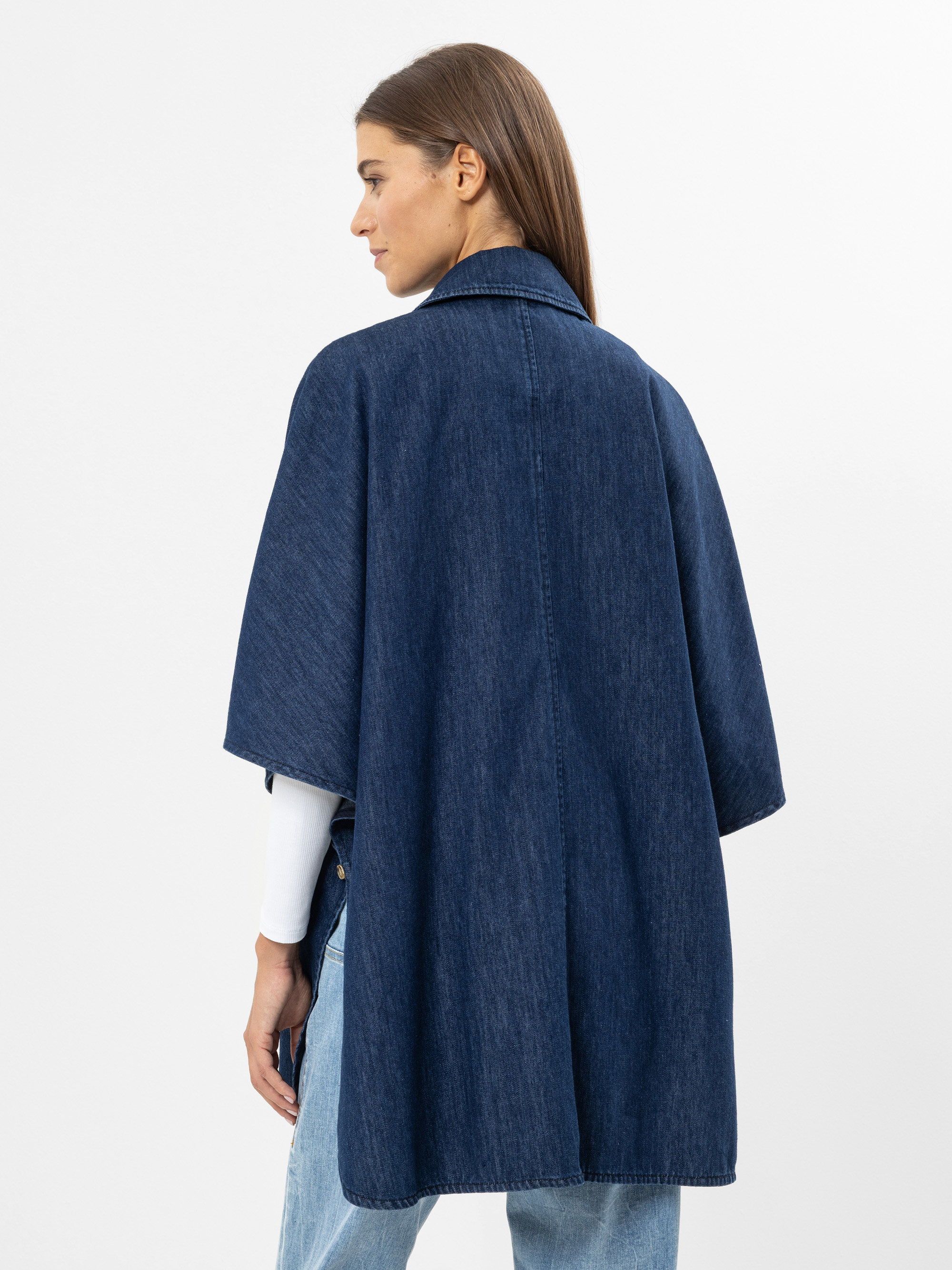 Eine Frau mit glattem braunem Haar, von hinten gesehen, trägt einen dunkelblauen Jeansumhang über einem weißen langärmeligen Oberteil und hellblaue Jeans vor einem schlichten weißen Hintergrund.