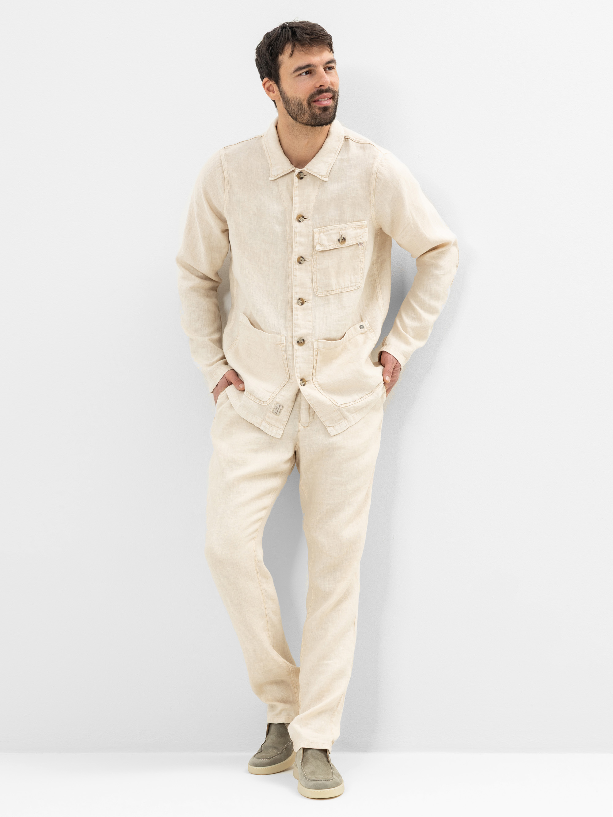 Ein Mann steht vor einem weißen Hintergrund und trägt eine hellbeige Button-up-Jacke mit mehreren Taschen und eine passende Hose. Er hat kurzes dunkles Haar und einen Bart und trägt olivgrüne Slipper. Seine Hände stecken in den Taschen.