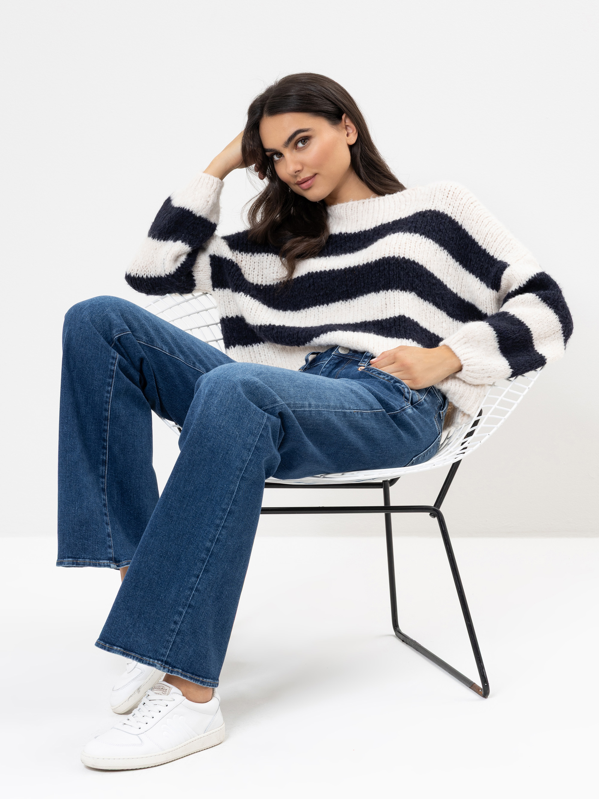 Eine Frau sitzt auf einem weißen Drahtstuhl und trägt einen weißen Pullover mit auffälligen schwarzen Streifen, blaue Jeans und weiße Turnschuhe. Sie hat dunkles, gewelltes Haar und stützt einen Ellbogen auf die Armlehne des Stuhls, während sie sel