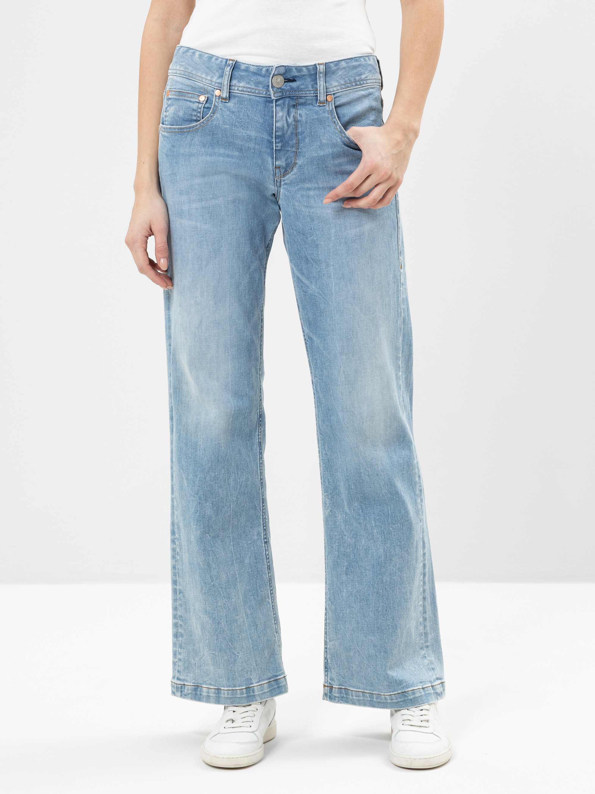 Eine Person, die hellblaue, weit geschnittene Jeans, ein weißes Oberteil und weiße Turnschuhe trägt, steht vor einem schlichten, hellen Hintergrund. Das Bild ist von den Schultern bis zu den Füßen abgeschnitten.