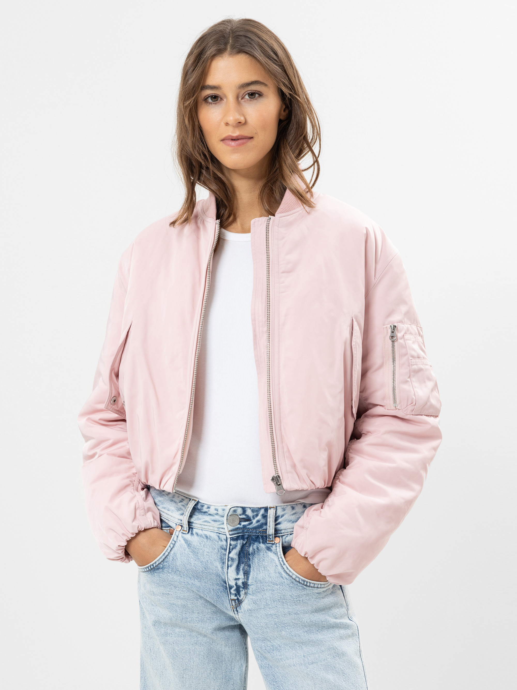 Eine Person, die eine hellrosa Bomberjacke über einem weißen Hemd und hellblaue Jeans trägt, steht mit den Händen in den Taschen vor einem schlichten weißen Hintergrund.