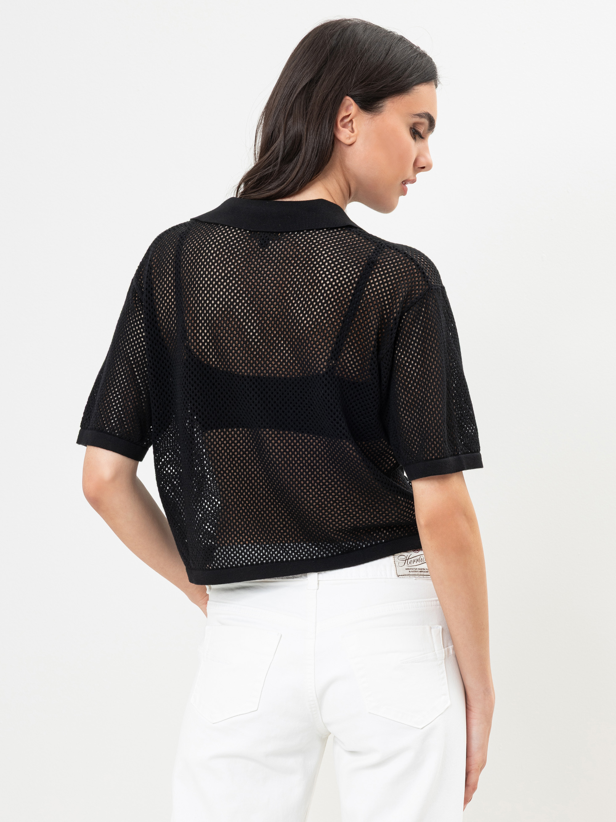 Eine Frau mit dunklem Haar trägt ein schwarzes, kurzärmeliges Netzoberteil über einem schwarzen BH und weiße Jeans. Sie steht mit dem Rücken zur Kamera vor einem schlichten, hellen Hintergrund.