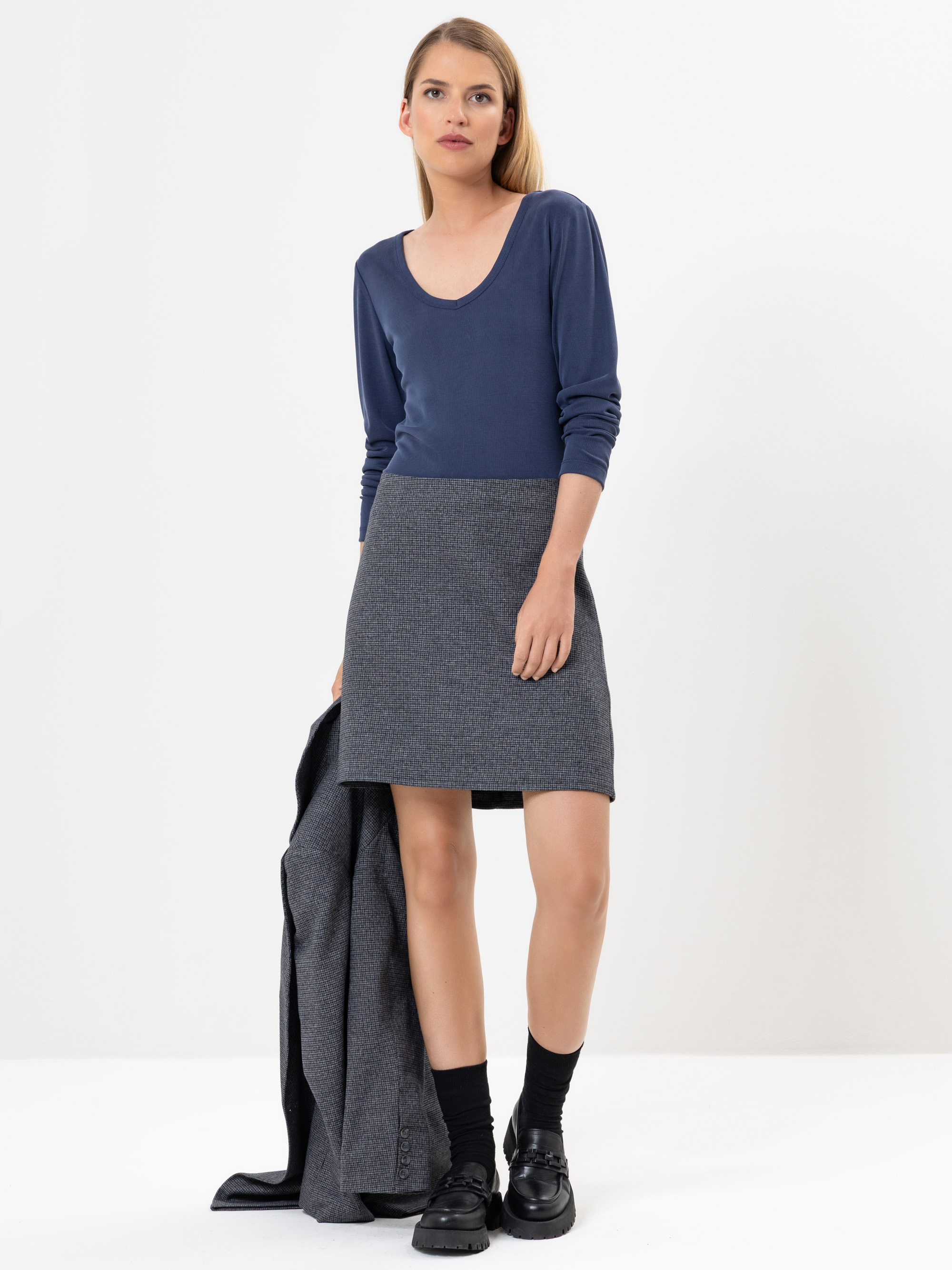Eine Frau mit glattem blondem Haar trägt ein langärmeliges marineblaues Oberteil und einen grauen Rock und hält einen grauen Mantel in einer Hand. Sie steht vor einem schlichten weißen Hintergrund und trägt schwarze Socken und schwarze Schuhe.