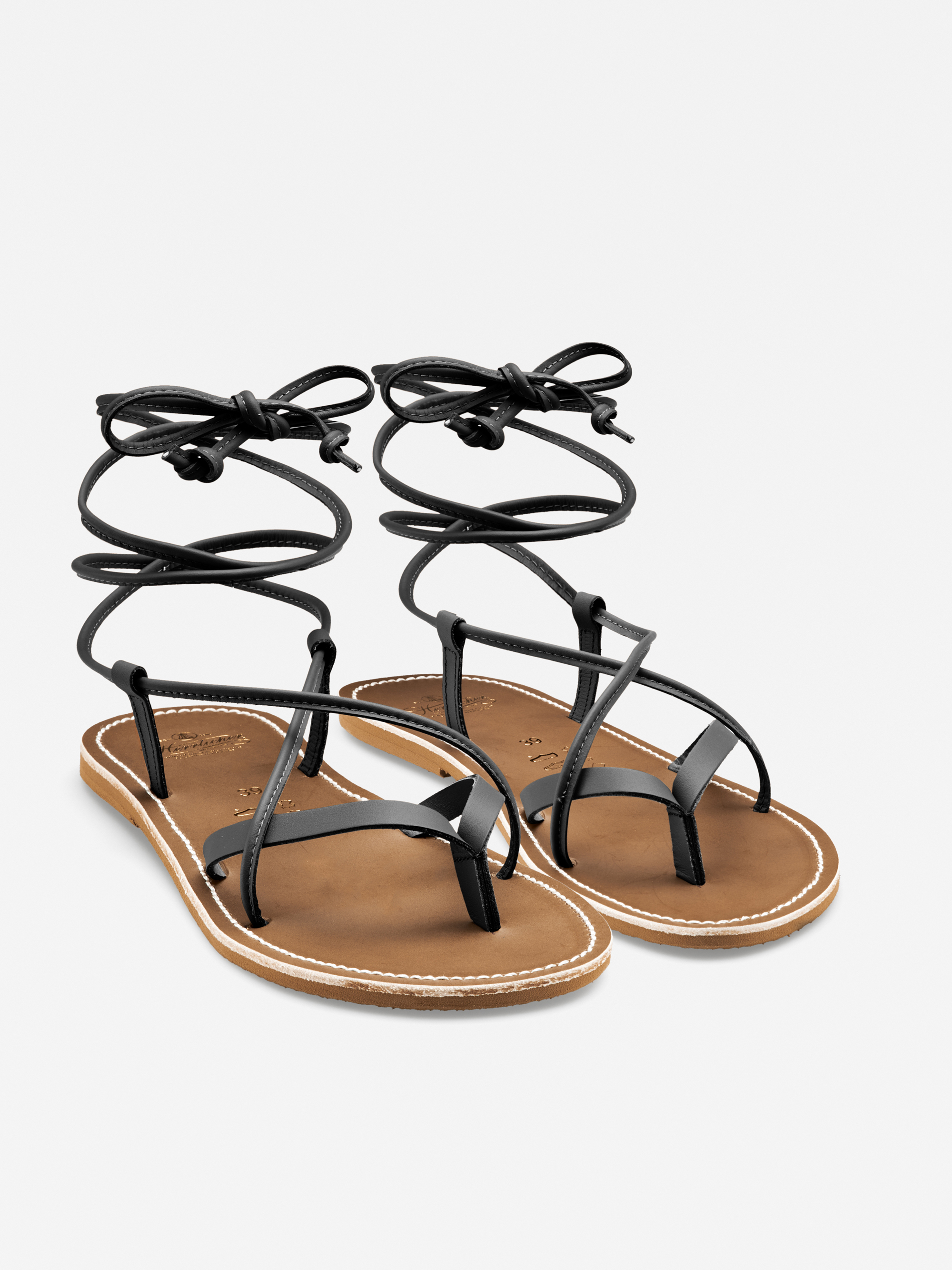 Ein Paar flache Sandalen mit hellbrauner Sohle, weißen Nähten und dünnen schwarzen Lederriemen, die um den Knöchel gewickelt und vorne mit einer Schleife gebunden werden.