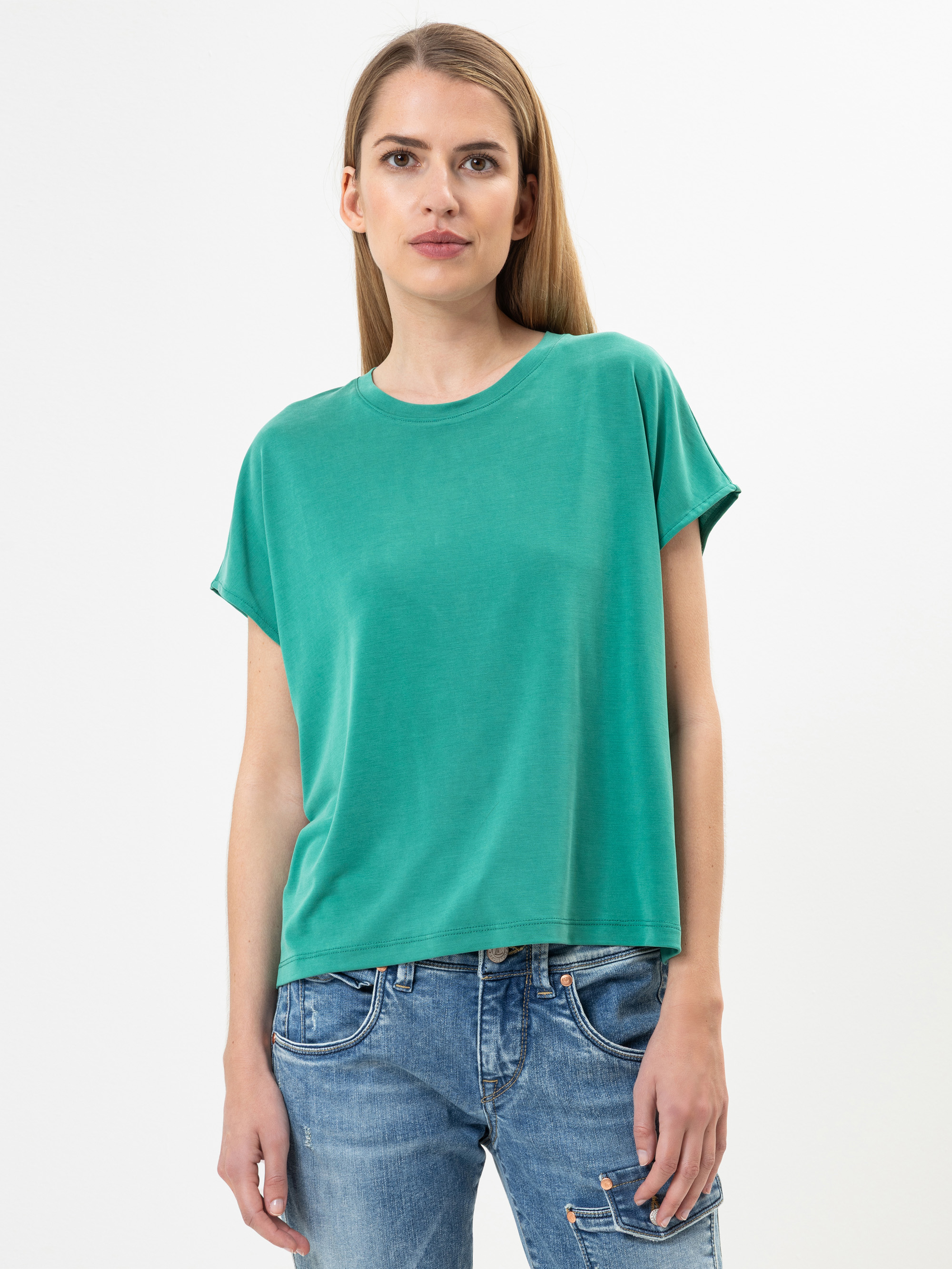 Eine Frau mit glattem, hellbraunem Haar, die ein lockeres grünes T-Shirt und eine blaue Jeans trägt, steht vor einem schlichten weißen Hintergrund und schaut mit neutralem Blick direkt in die Kamera.