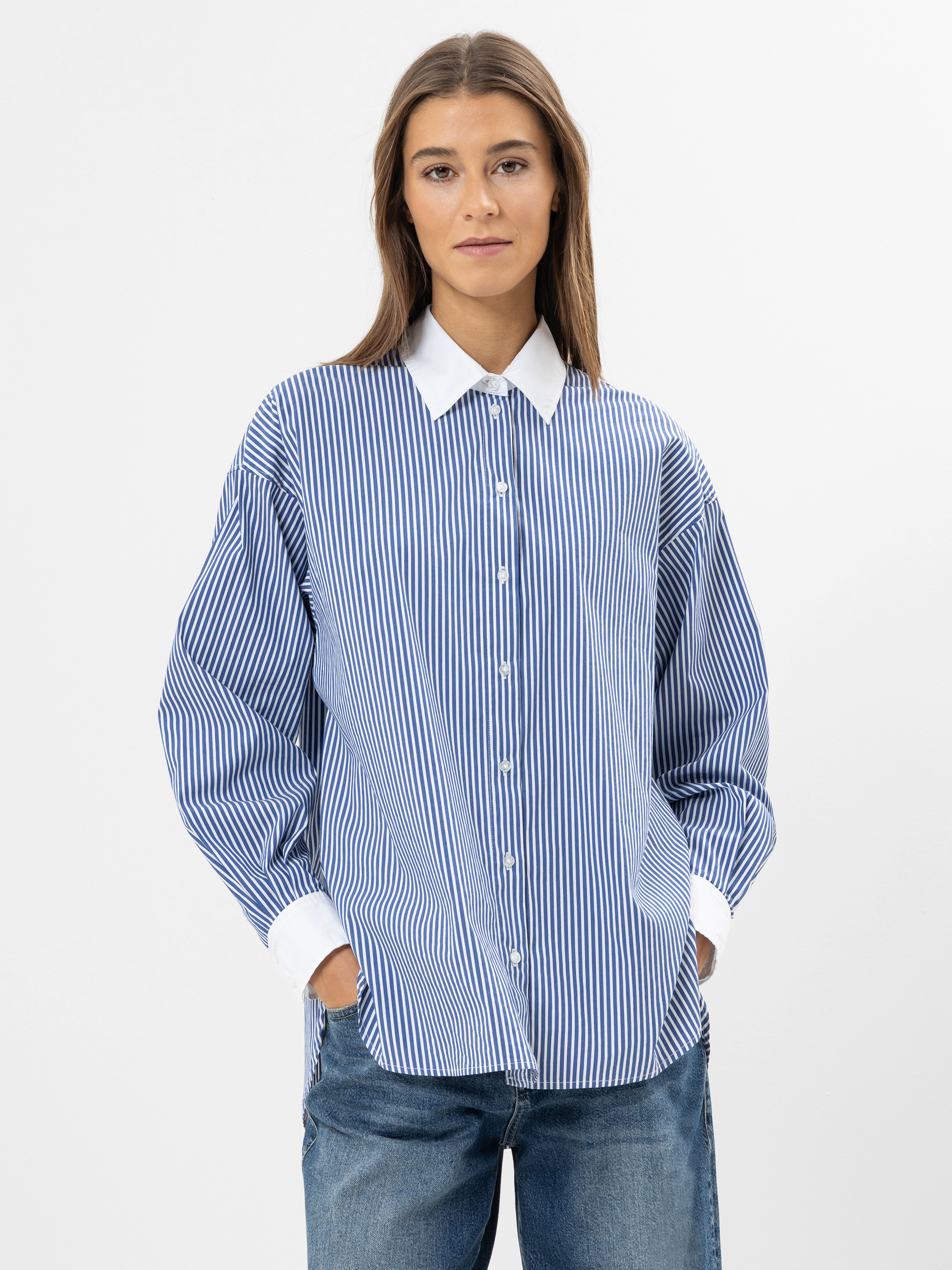 Eine Frau, die ein blau-weiß gestreiftes Button-up-Hemd mit weißem Kragen und Manschetten und eine blaue Jeans trägt, steht vor einem schlichten weißen Hintergrund und hat die Hände in den Taschen.