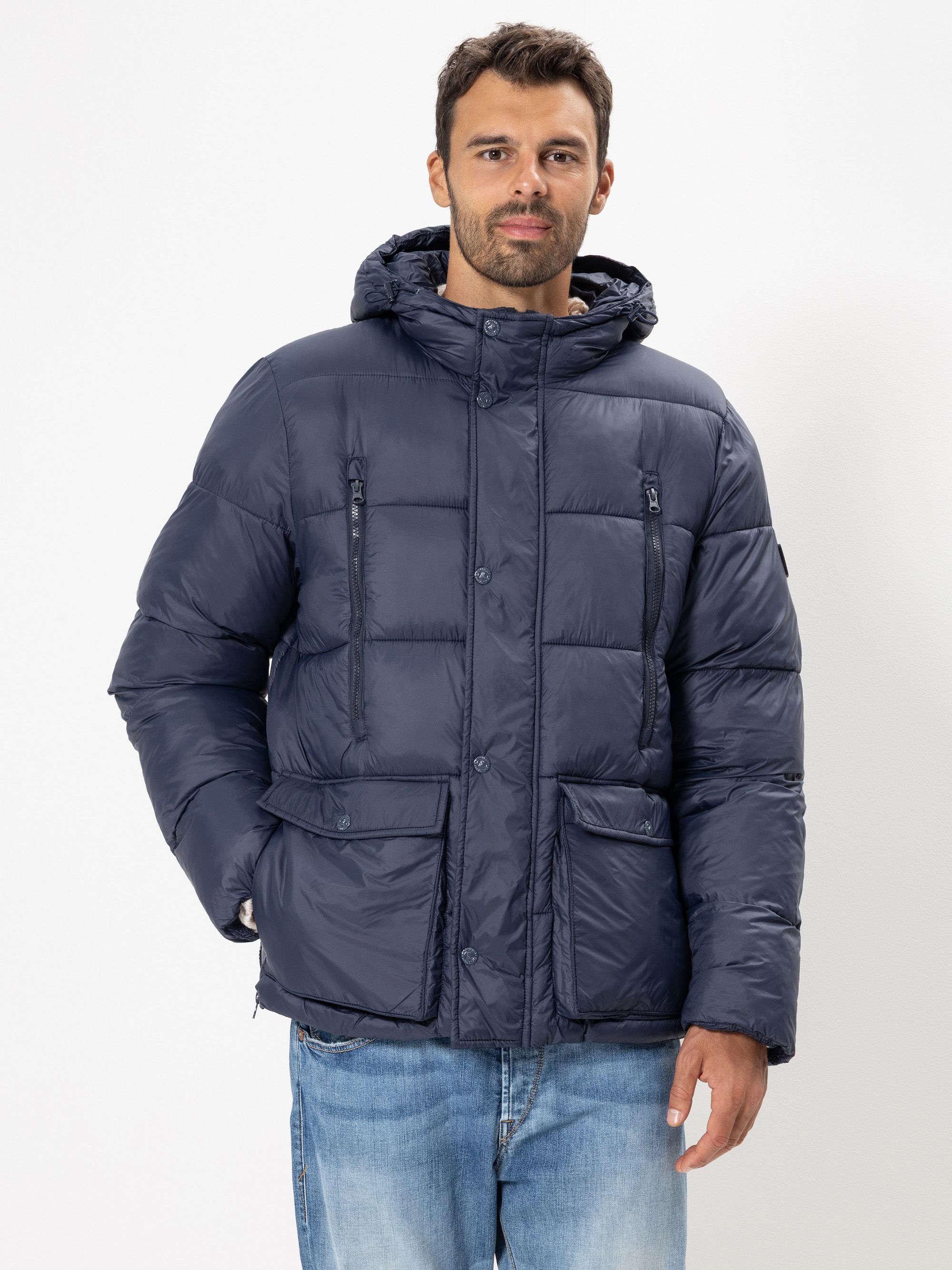 Ein Mann mit kurzen dunklen Haaren und Bart trägt eine marineblaue Pufferjacke mit Kapuze und blaue Jeans und steht vor einem schlichten weißen Hintergrund.