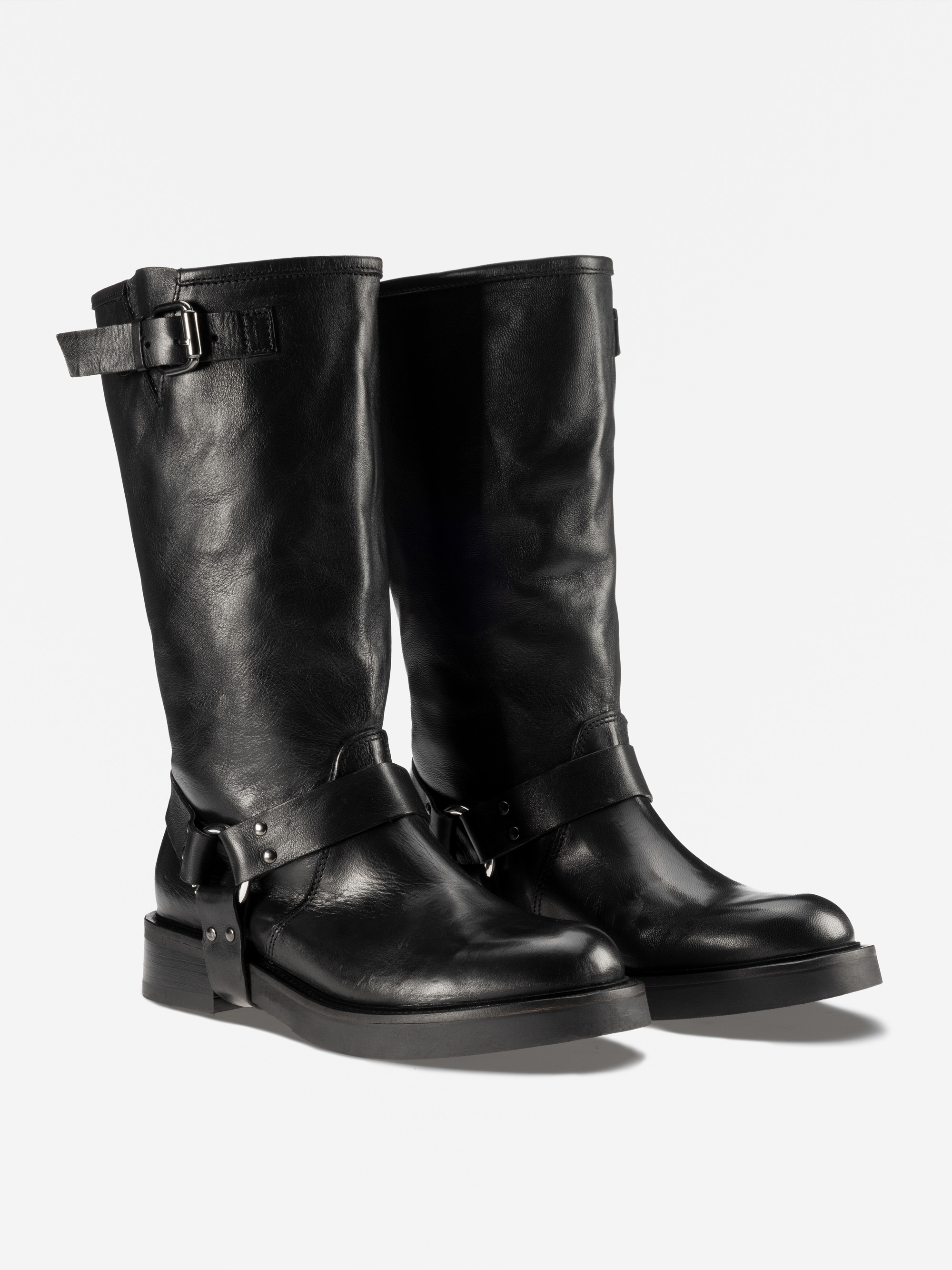 Ein Paar hoher schwarzer Lederstiefel mit niedrigem Absatz, dekorativen Schnallenriemen um den Knöchel und oben und einer leicht abgerundeten Spitze, dargestellt auf einem schlichten weißen Hintergrund.