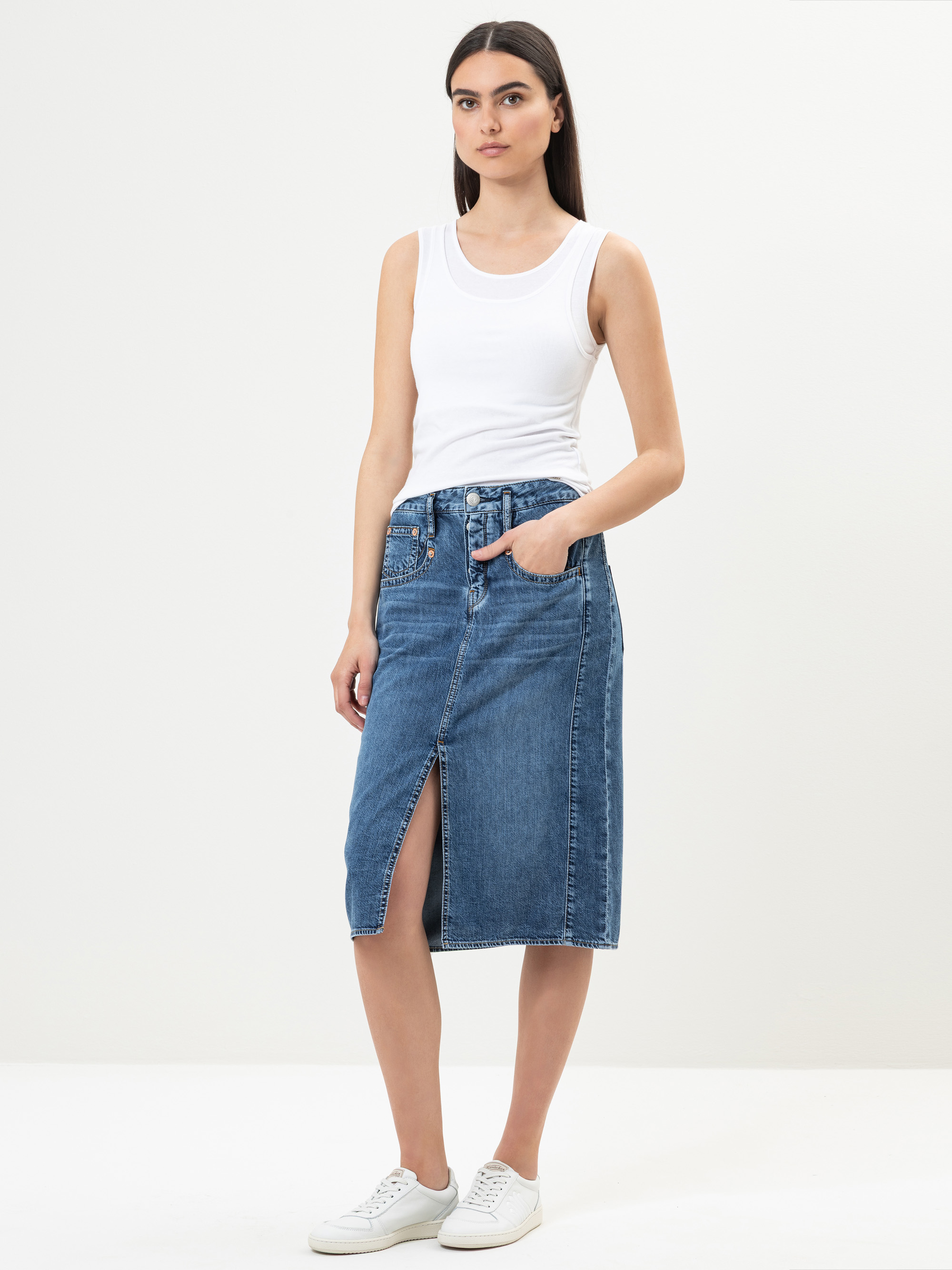 Eine Frau steht vor einem unifarbenen Hintergrund und trägt ein weißes ärmelloses Oberteil, einen knielangen blauen Jeansrock mit einem Schlitz vorne und weiße Turnschuhe. Sie hat langes, glattes dunkles Haar und hat die Hände in den Rocktaschen.