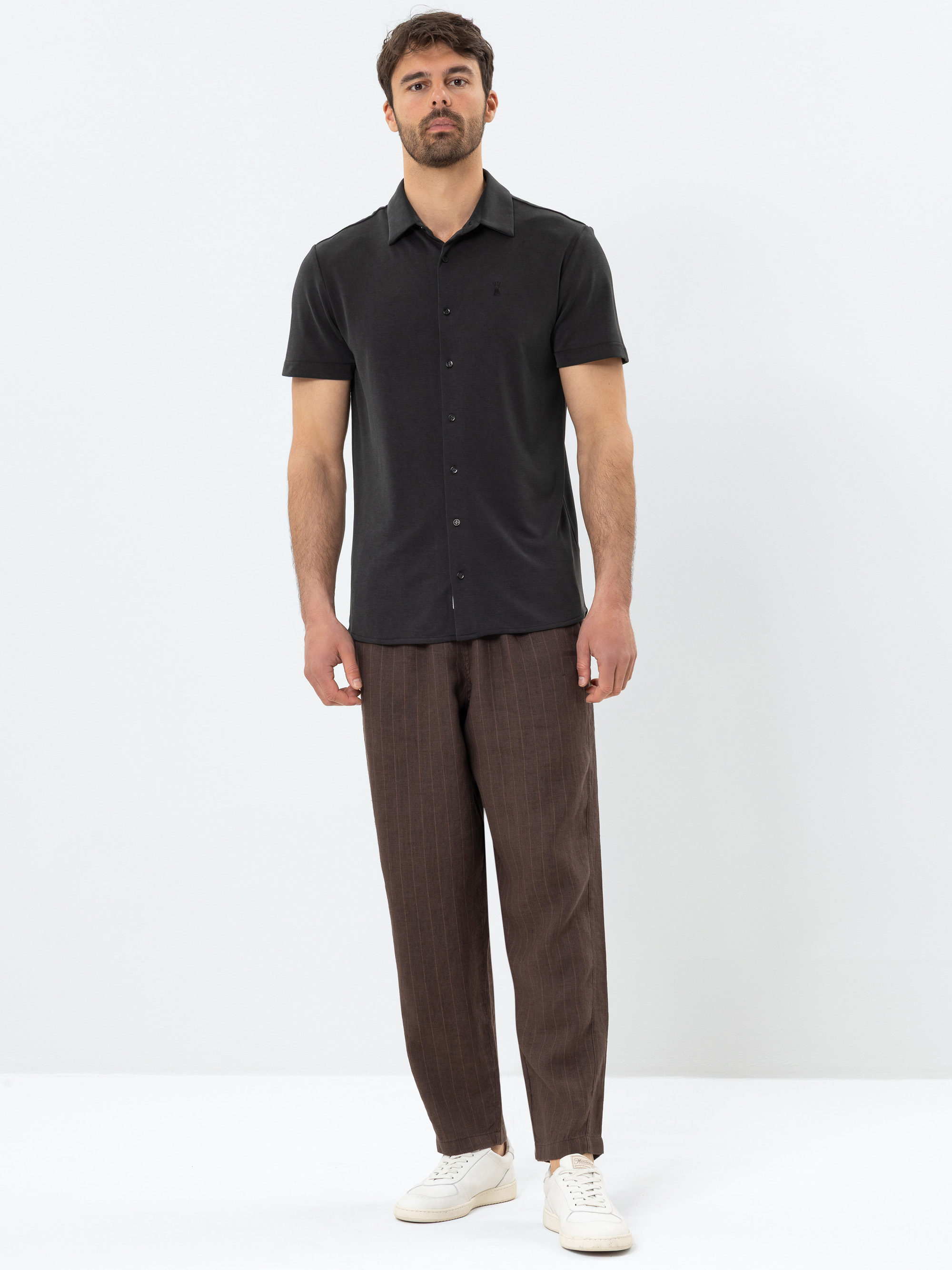 Ein Mann steht vor einem schlichten weißen Hintergrund und trägt ein kurzärmeliges schwarzes Button-up-Hemd, eine weite braune Hose und weiße Turnschuhe. Er hat kurzes dunkles Haar und einen Bart, hat einen neutralen Gesichtsausdruck und eine entspann