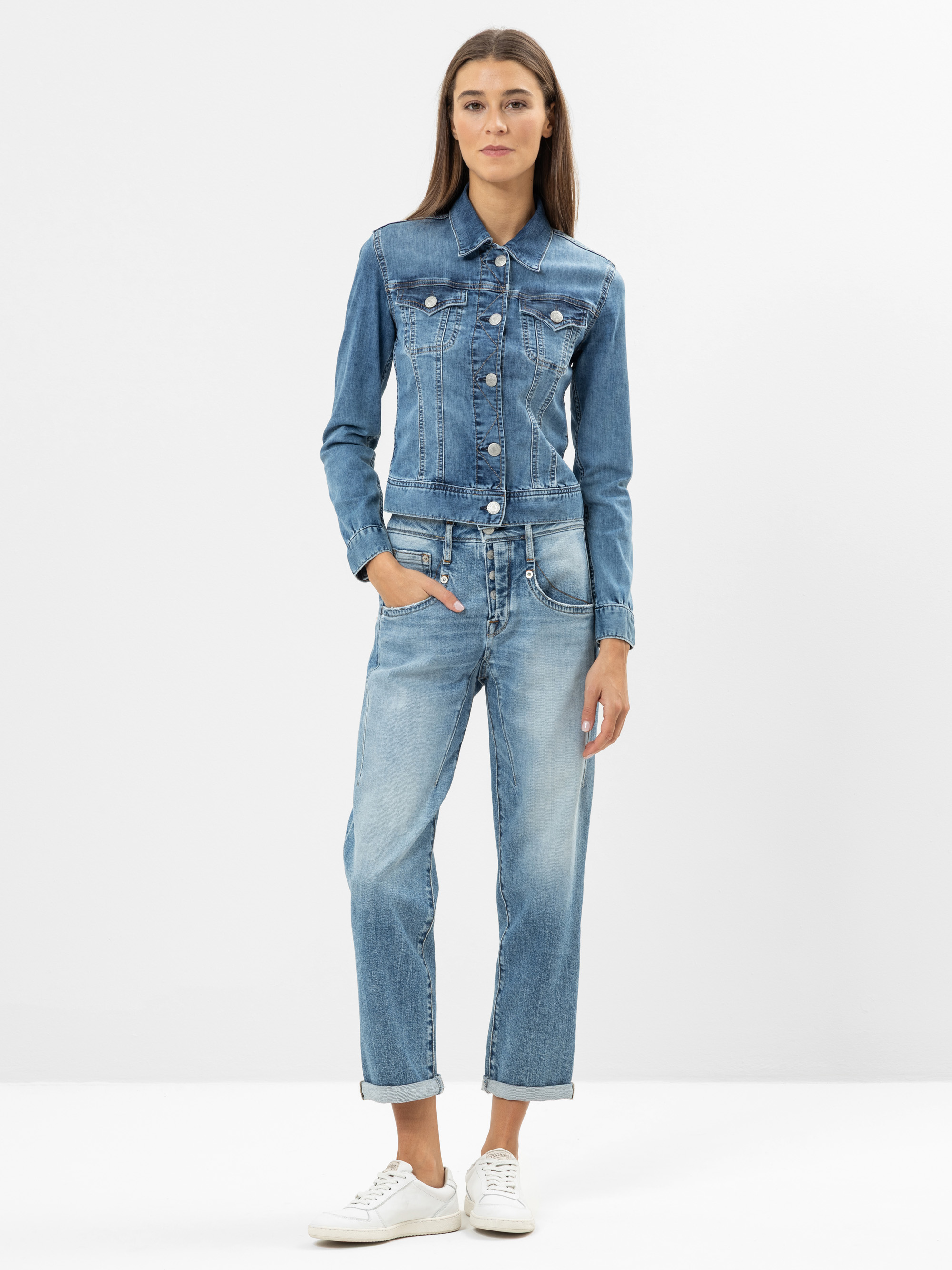 Eine Frau steht vor einem schlichten weißen Hintergrund und trägt eine blaue Jeansjacke, dazu passende Jeans und weiße Turnschuhe. Sie hat langes braunes Haar und ihre Hände stecken in den Taschen.