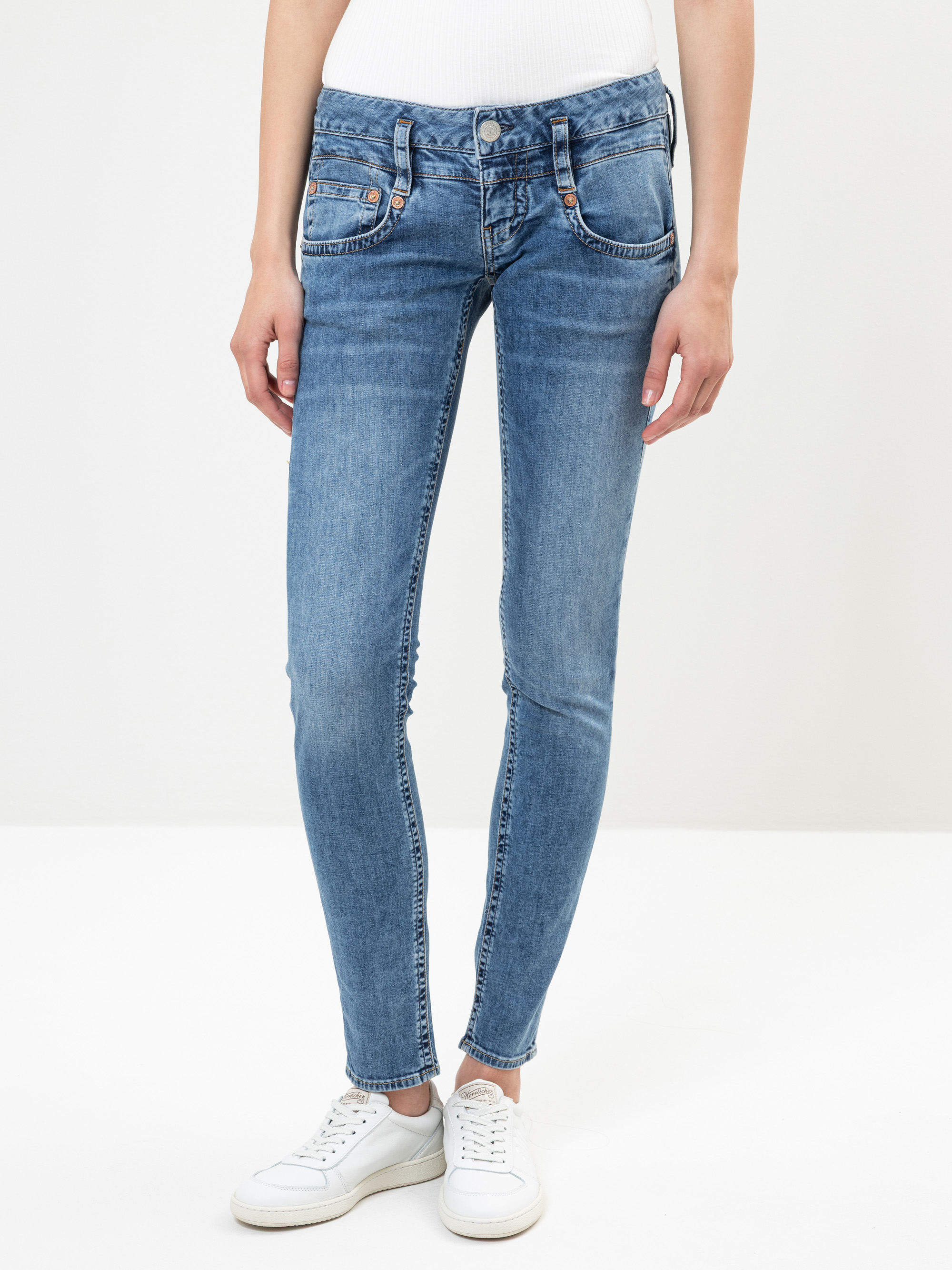 Eine Person in hellblauen Skinny-Jeans und weißen Turnschuhen steht vor einem schlichten weißen Hintergrund. Nur der untere Teil des Oberkörpers und die Beine sind sichtbar.