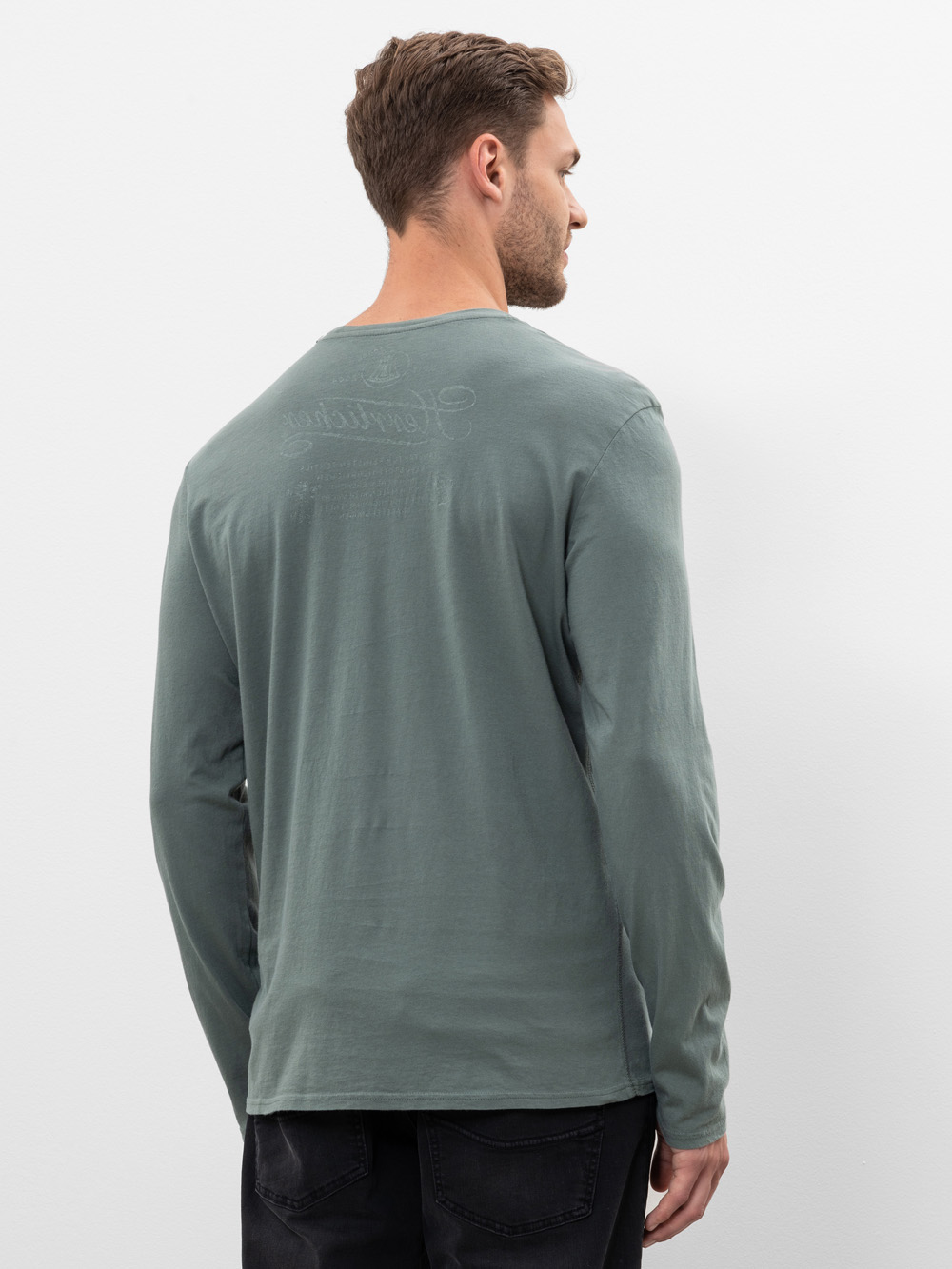 Ein Mann mit kurzen braunen Haaren steht mit dem Rücken zur Kamera, trägt ein langärmeliges, gedämpftes grünes Hemd und schwarze Jeans vor einem schlichten weißen Hintergrund.