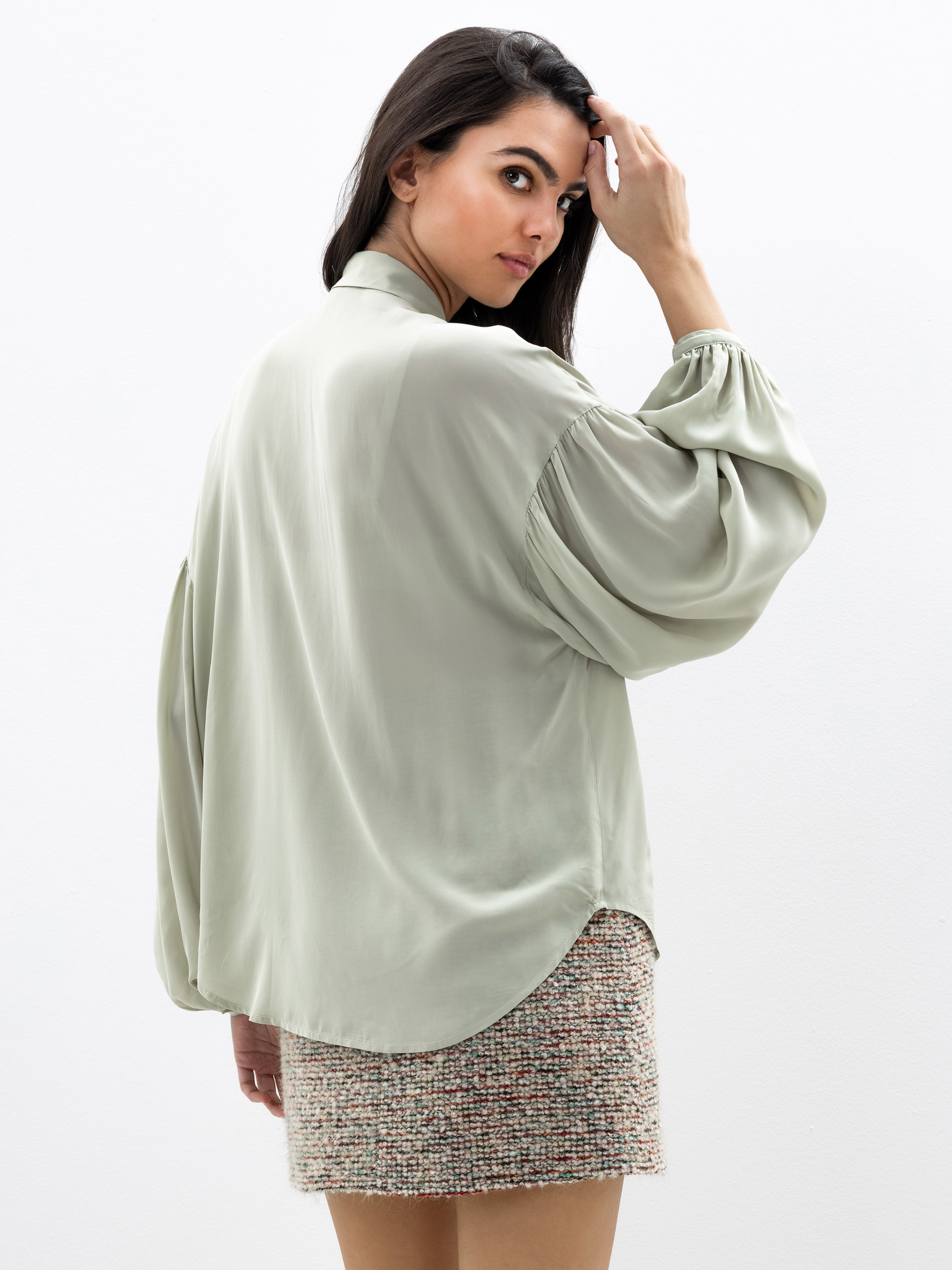 Eine Frau steht leicht abgewandt und schaut über ihre Schulter. Sie trägt eine lockere, hellgrüne Bluse mit weiten Ärmeln und einen strukturierten, kurzen Tweed-Rock. Der Hintergrund ist schlicht und weiß.