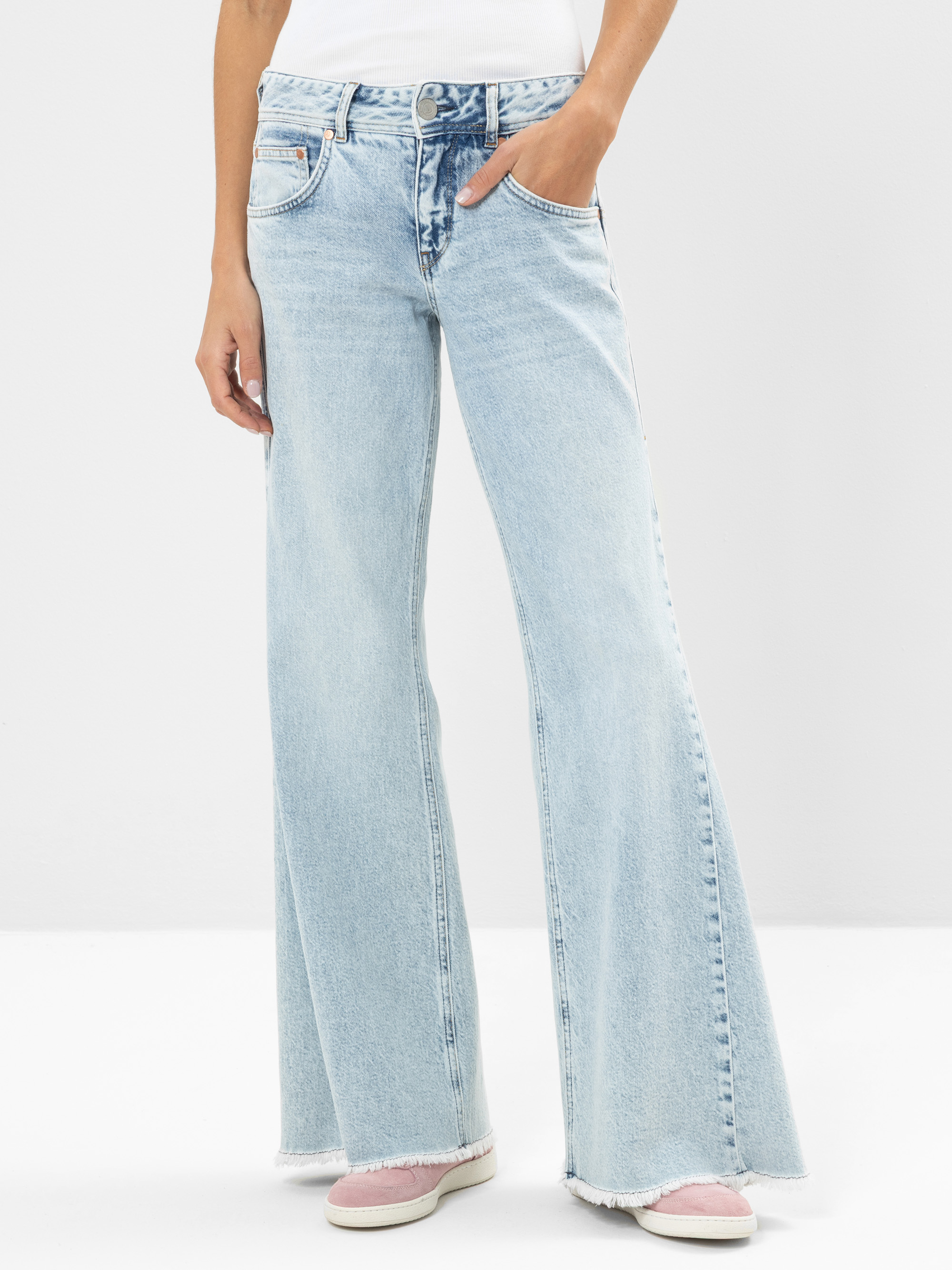 Eine Person, die hellblaue Jeans mit hoher Taille und ausgefransten Säumen, ein weißes Oberteil und rosa Turnschuhe trägt, steht vor einem schlichten hellen Hintergrund.
