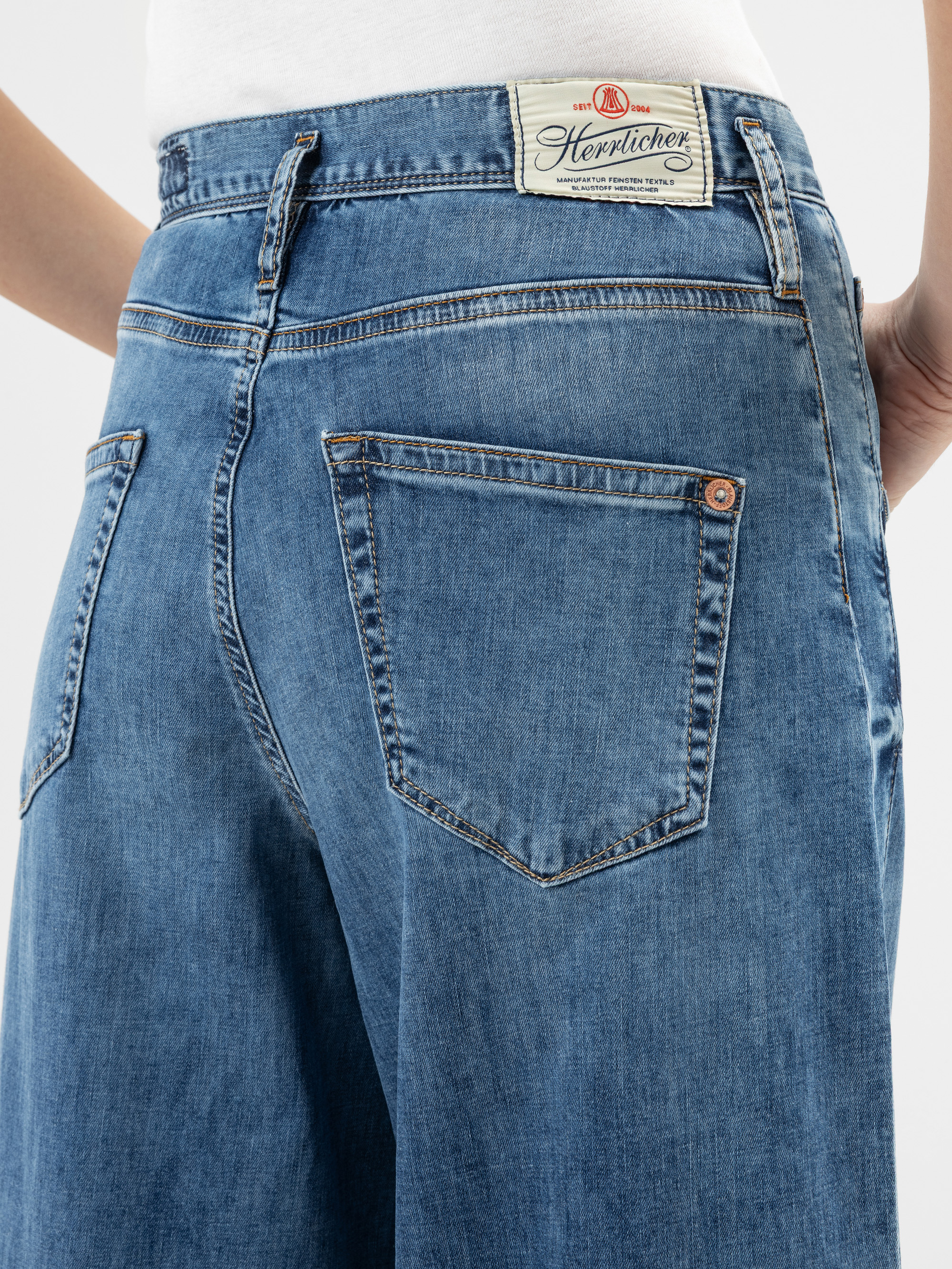 Nahaufnahme einer Person, die eine blaue Denim-Jeans trägt, mit der Gesäßtasche und einem weißen Herrlicher-Markenetikett am Bund. Die Person steht und hat die Hände an die Seite gelegt.