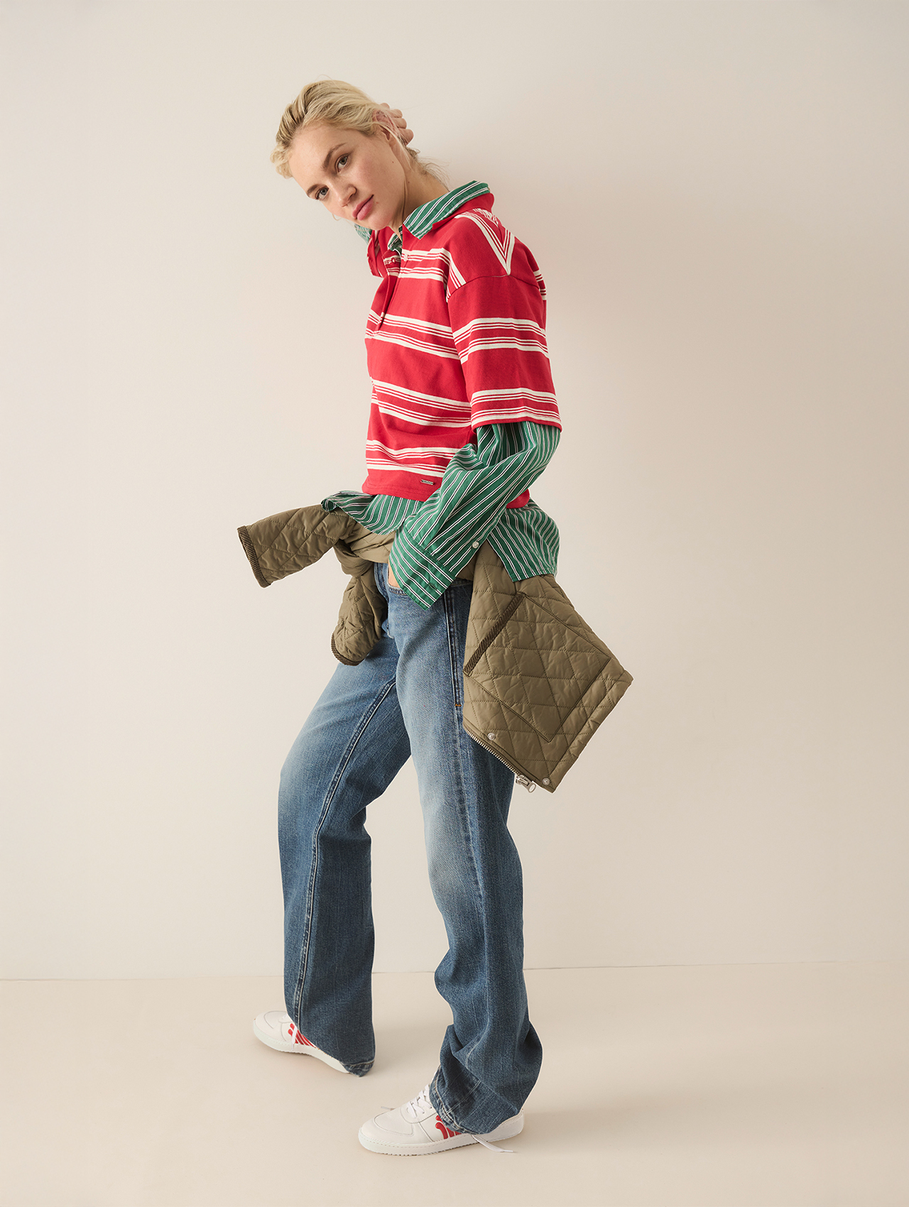 Eine Person steht vor einem schlichten, hellen Hintergrund und trägt ein rot gestreiftes Polohemd über einem grün gestreiften Hemd, blaue Jeans, weiße Turnschuhe und eine olivfarbene Steppjacke, die um die Taille gebunden ist. Sie blickt leicht zur Se